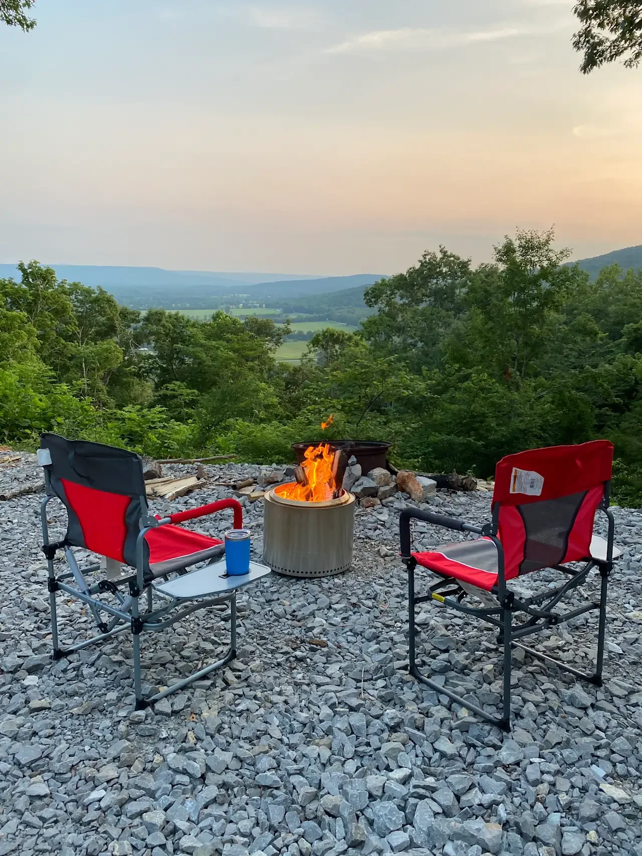 Two red and black camping chairs facing a fire pit on a rocky surface, overlooking a lush green landscape with trees and distant hills under a partly cloudy sky.