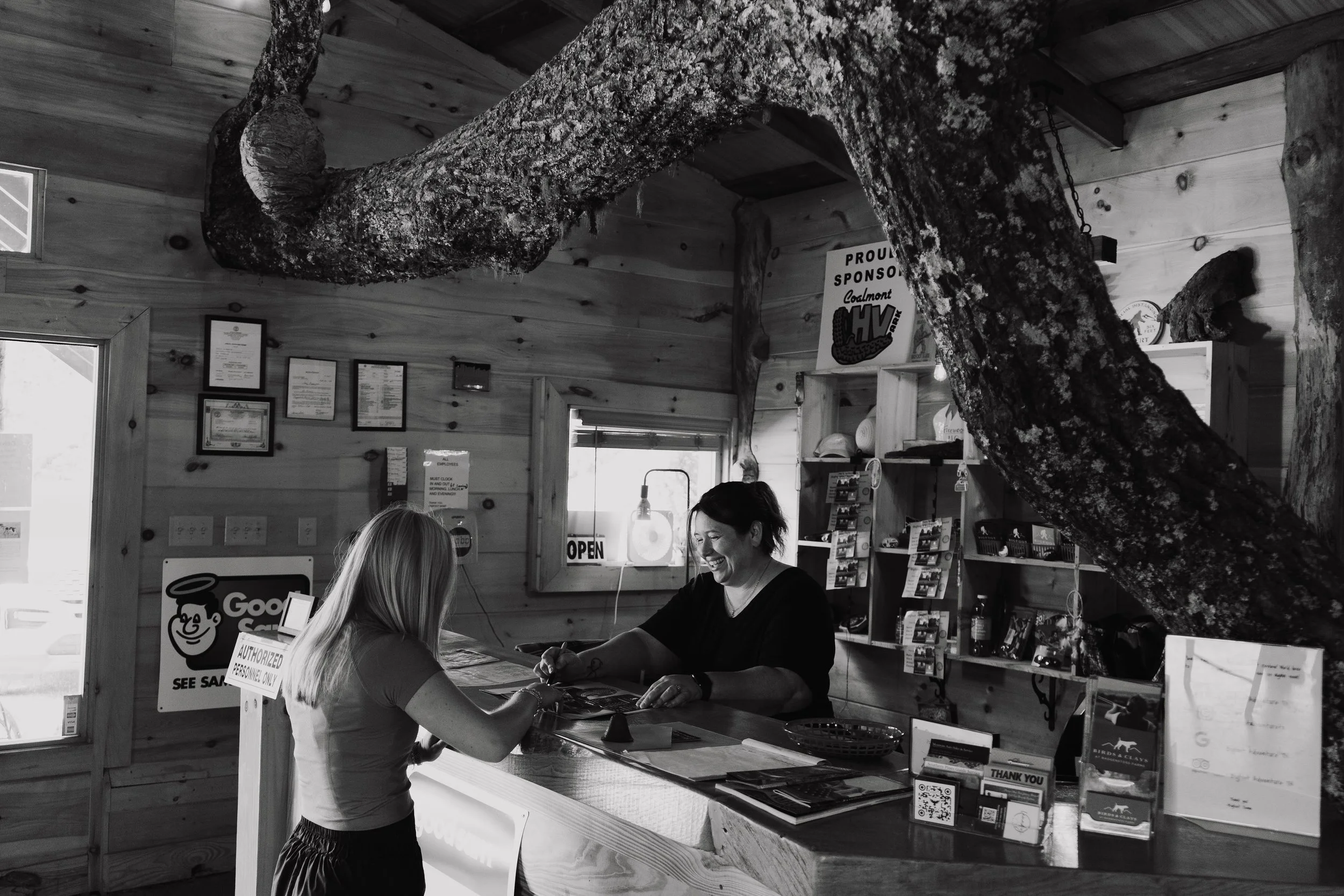 A smiling woman at a wooden counter interacting with a customer inside a rustic shop with wooden walls and shelves.