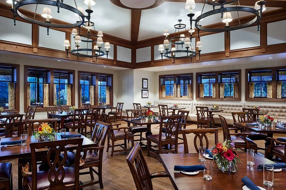 Elegant restaurant interior with wooden furniture, floral centerpieces, large windows, and chandeliers.