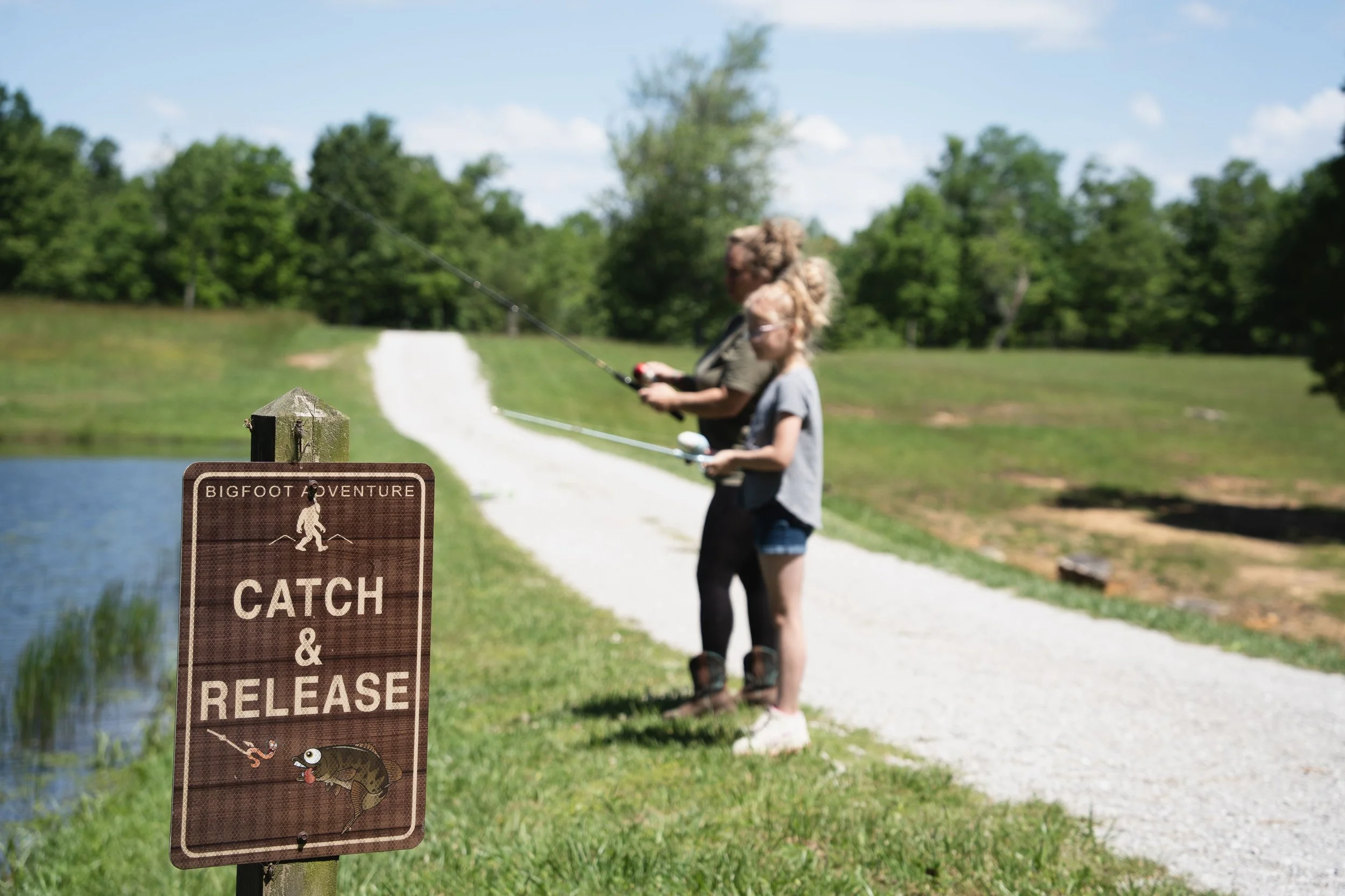 Two children fishing by a pond, standing on a gravel path, with a wooden sign that reads "Bigfoot Adventure, Catch & Release," and surrounded by greenery and trees.