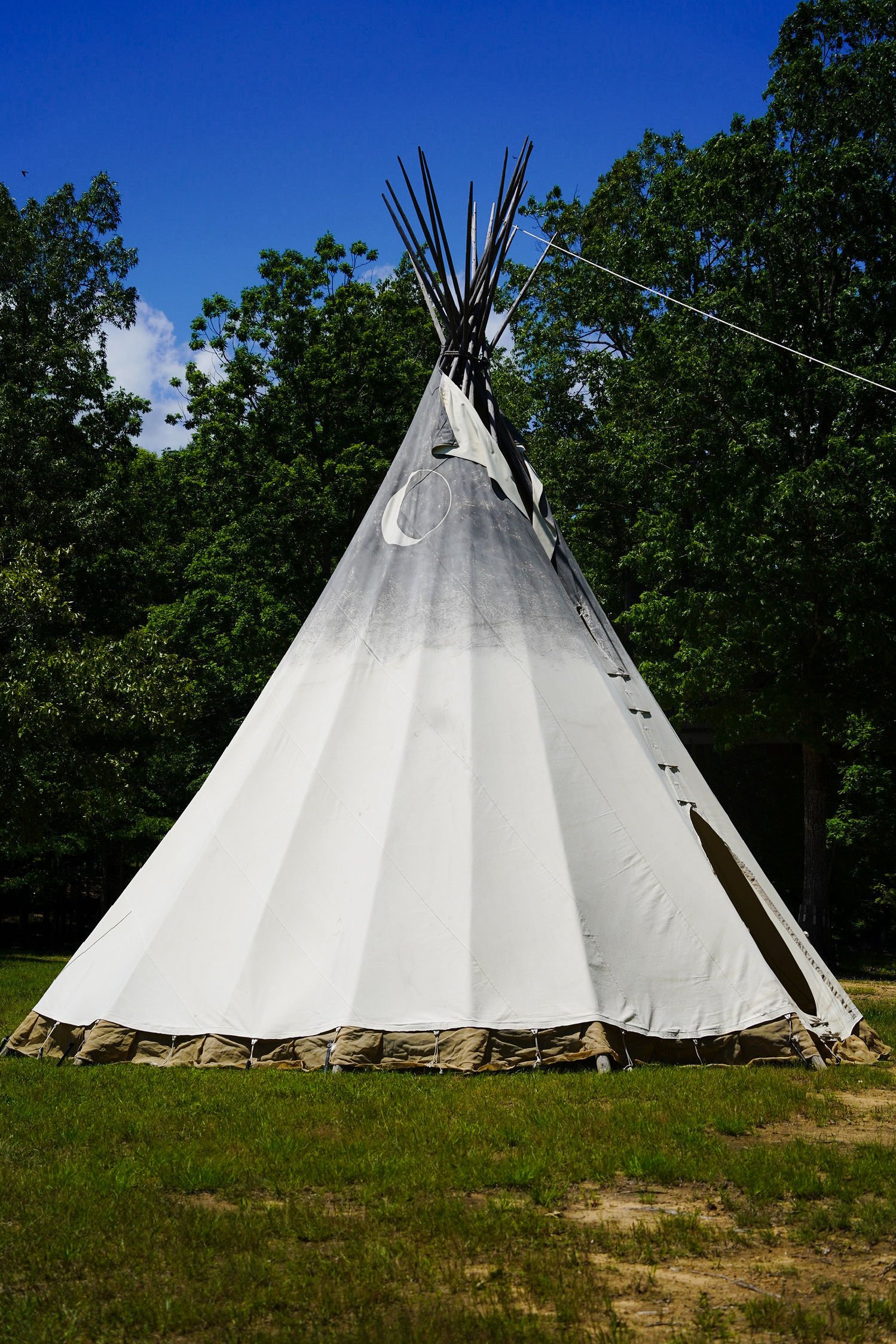 A traditional Native American teepee tent set up outdoors on grass with a backdrop of green trees and a partly cloudy blue sky.