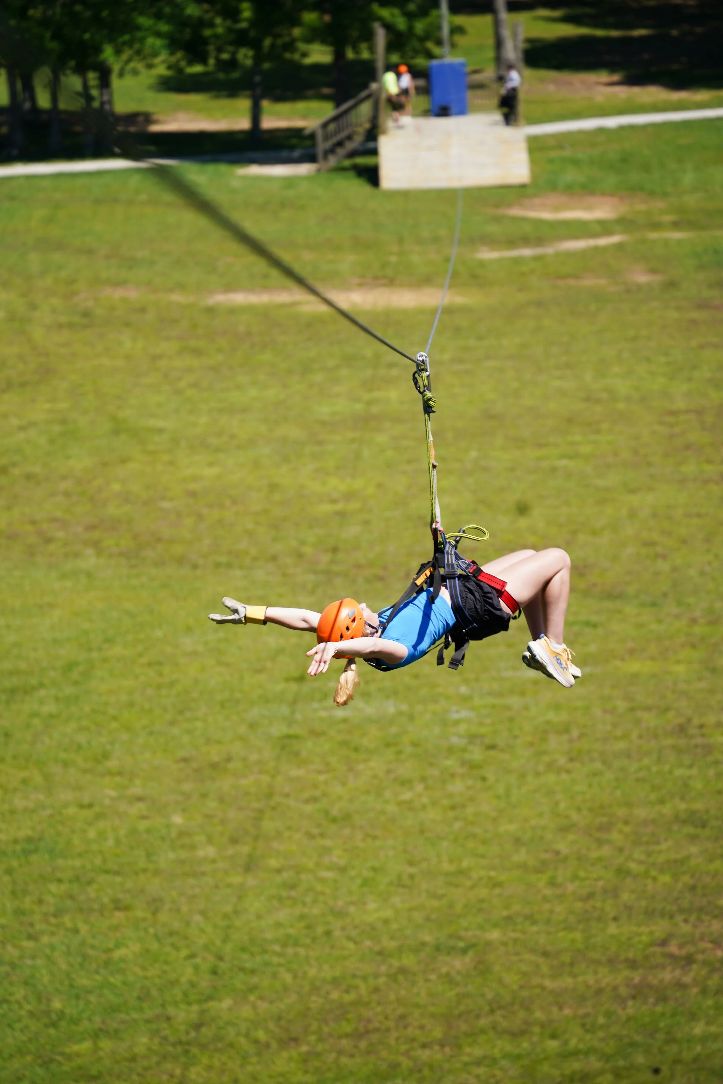 Child ziplining in a harness, wearing an orange helmet and gloves, with arms outstretched, over a grassy field.