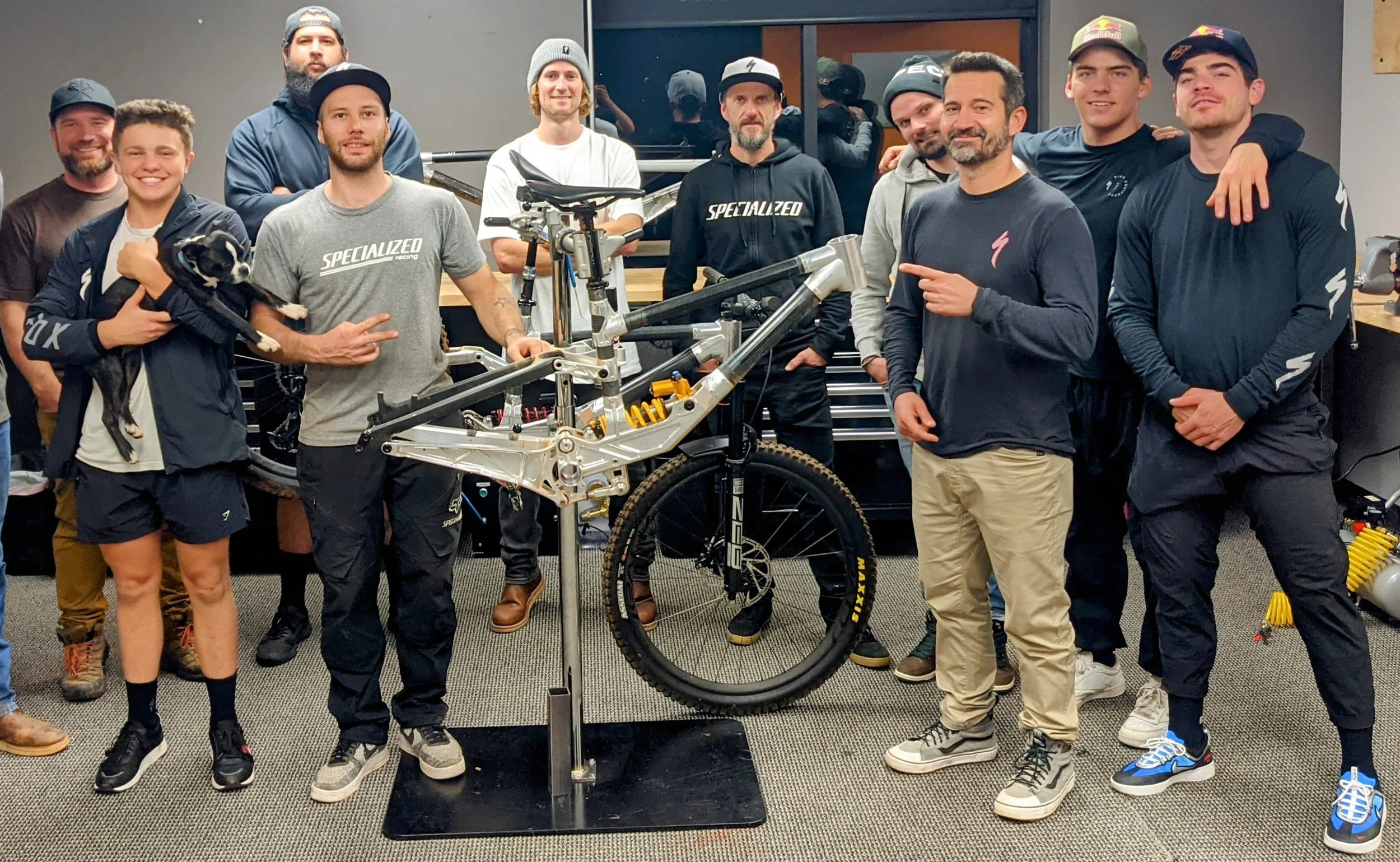 Group of men and a woman posing around a mountain bike in a workshop. Some are smiling and pointing at the bike, with a small dog being held by the woman.