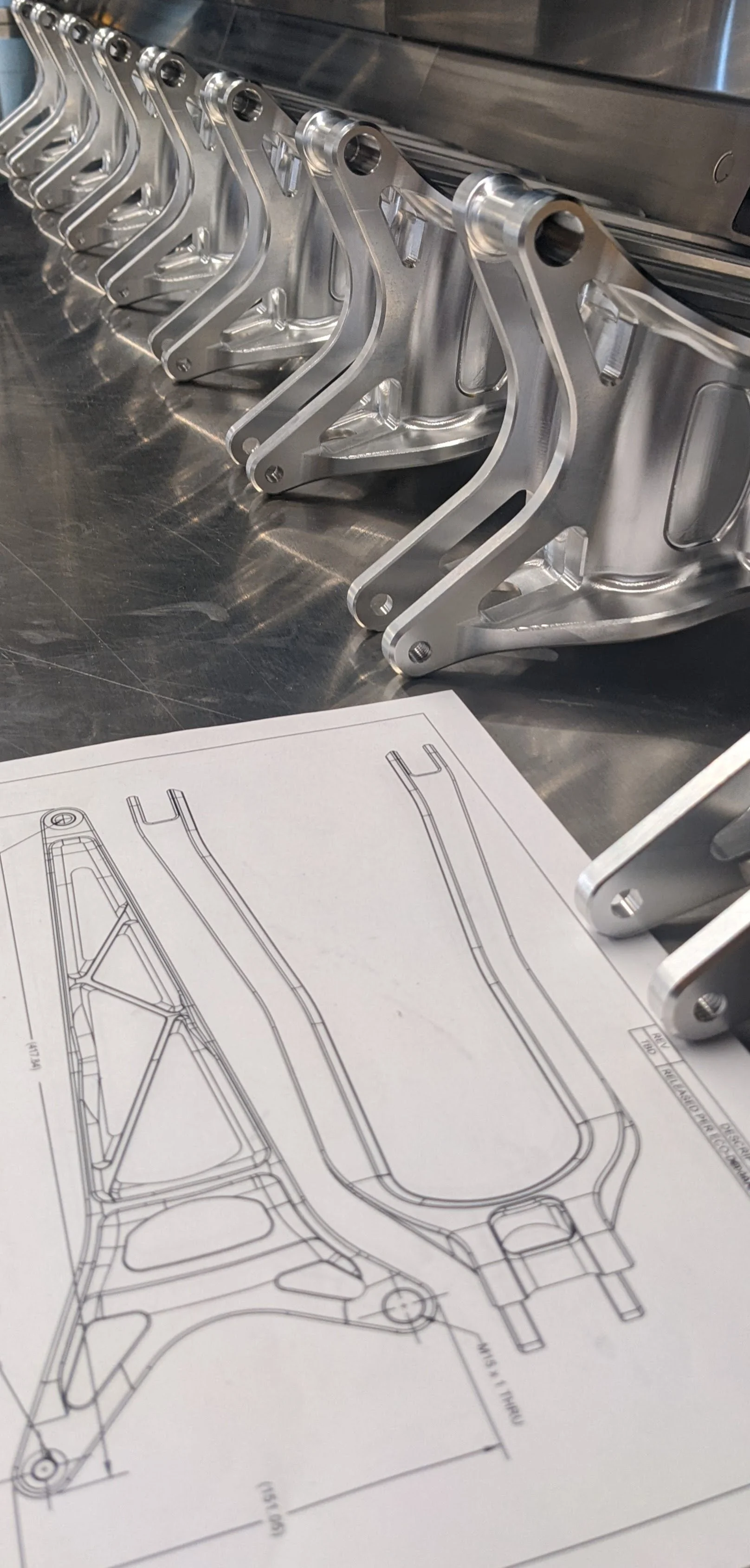 Multiple machined aluminum parts lined up on a work surface with a technical drawing underneath.