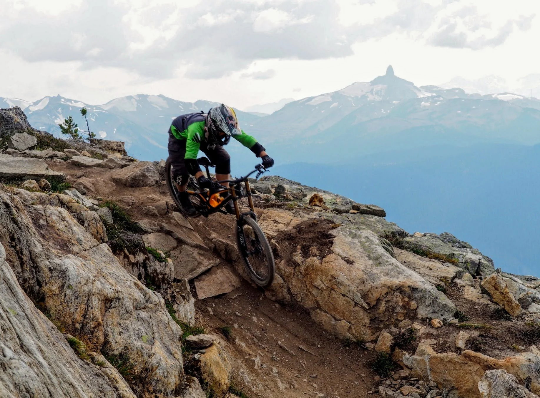 A mountain biker wearing a helmet and green and black gear navigates a rocky trail on a mountain slope, with a mountain range in the background. Location is top of the world in whistler.