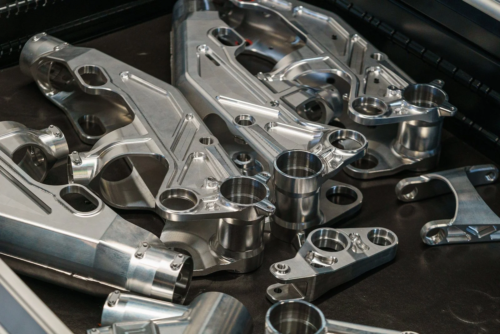 Collection of shiny, metallic mechanical parts with hollow cylindrical sections, complex curves, and bolt holes on a black surface.