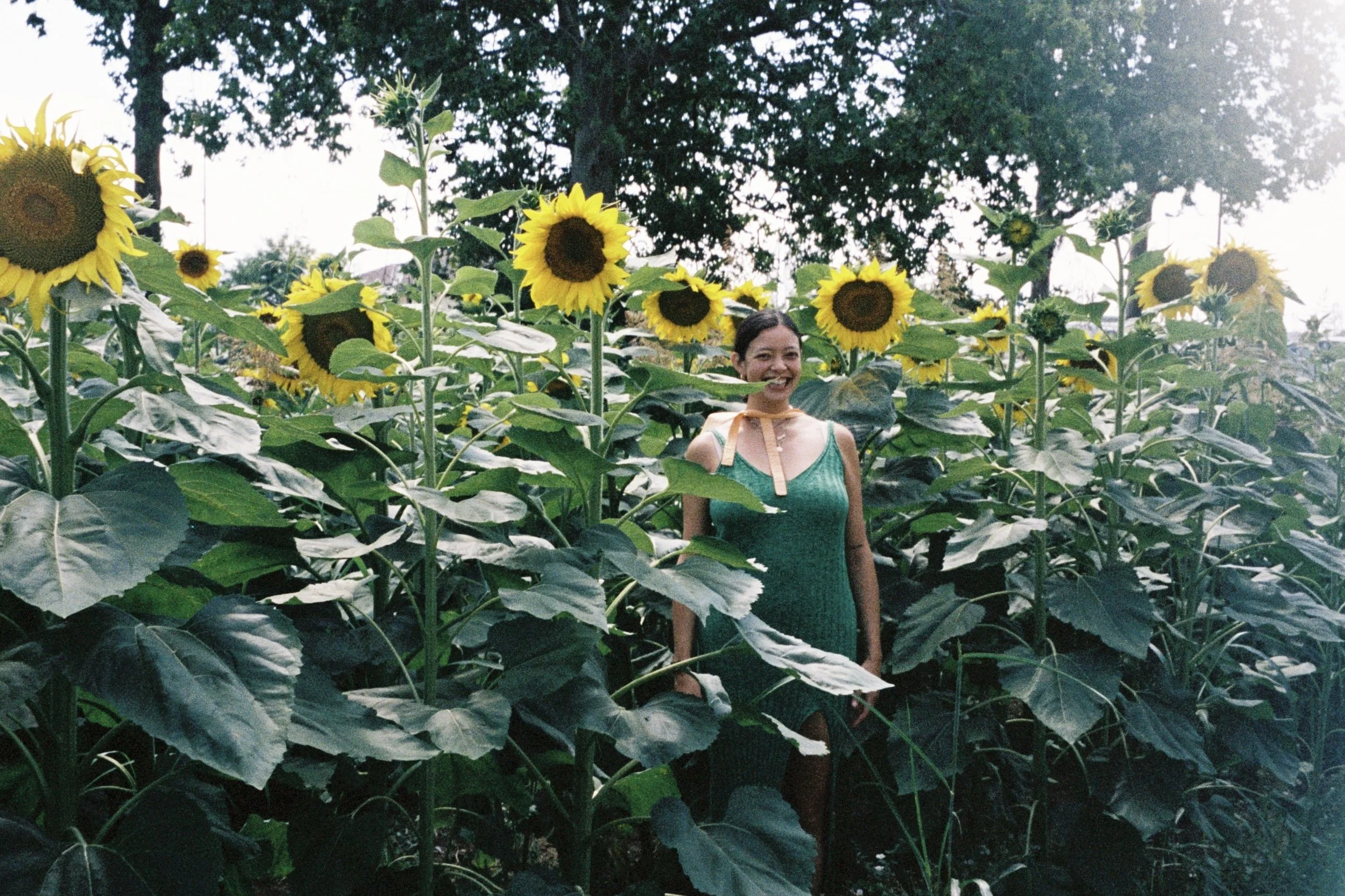 A woman in a green dress stands among tall sunflowers and smiles outdoors. There are trees and a cloudy sky in the background.