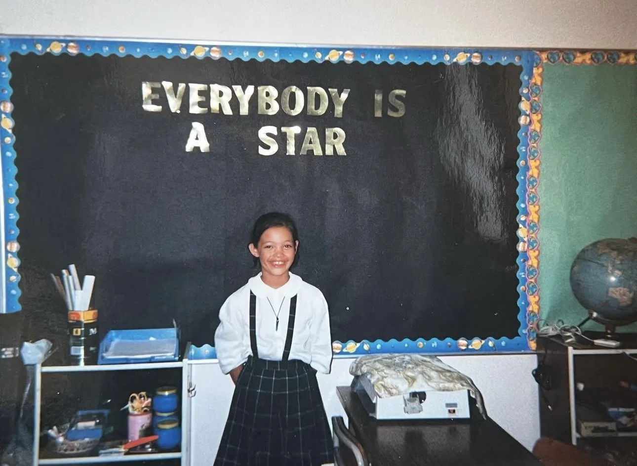A young girl stands smiling in front of a black chalkboard with a motivational quote that says, 'Everybody is a star,' in a classroom.