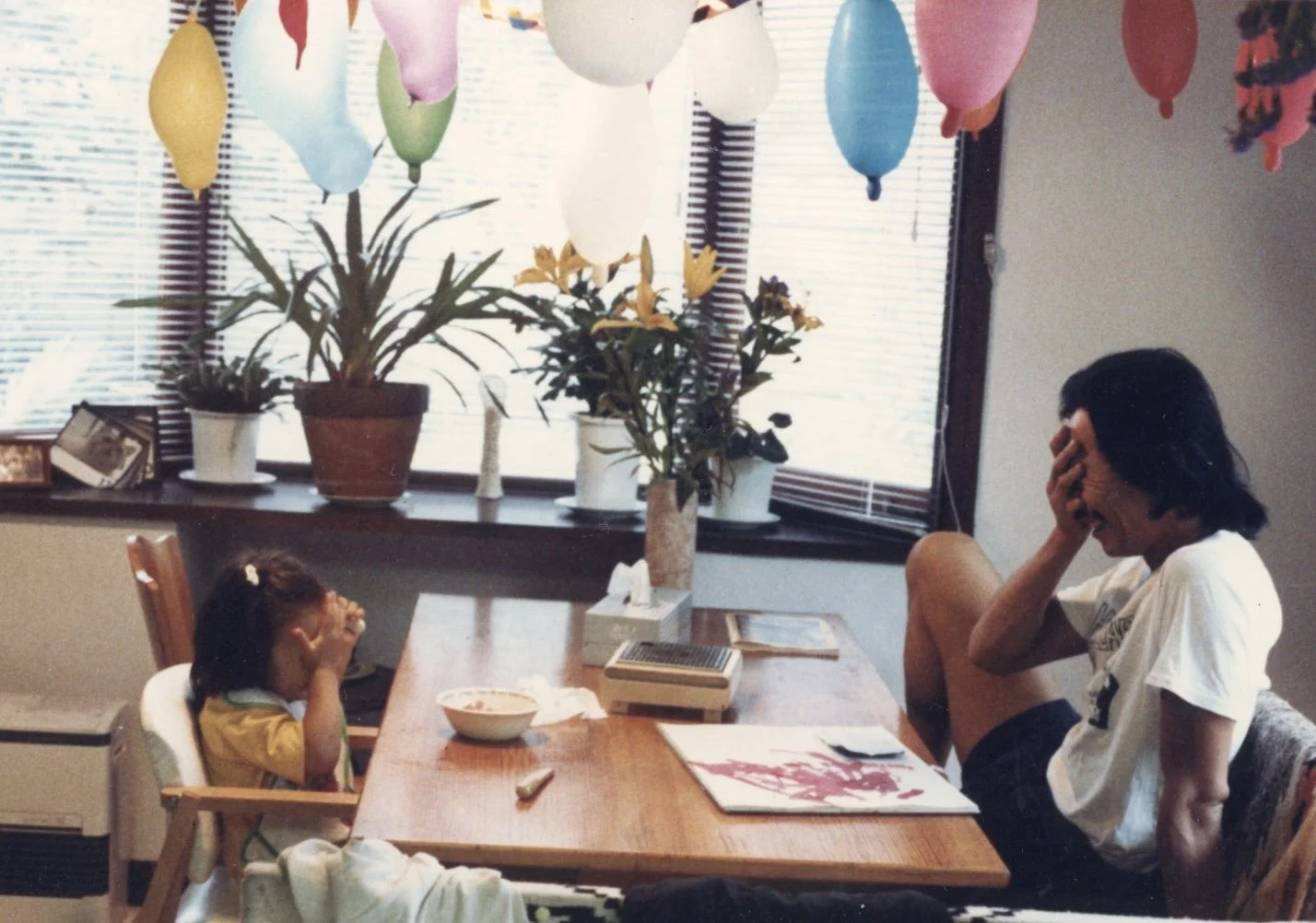 A man with dark hair and a young girl, both sitting at a wooden dining table, covering their faces in a gesture of laughter or shyness. The room is decorated for a celebration with colorful balloons hanging from the ceiling and potted plants on the window sill.