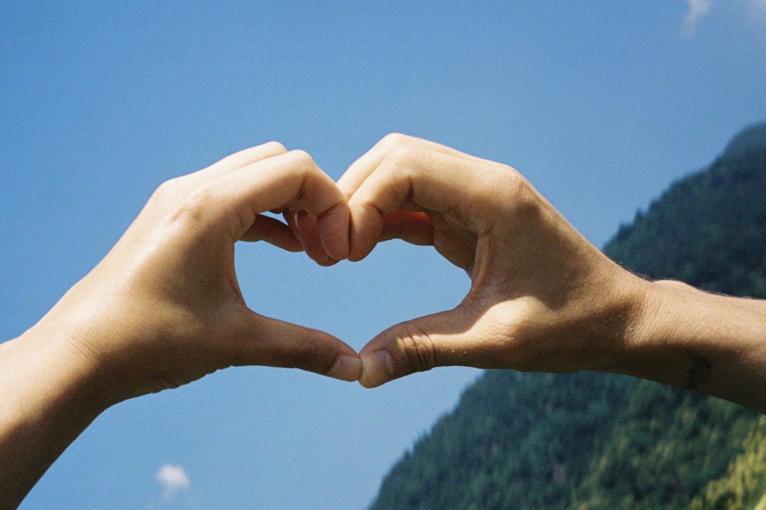 Two hands forming a heart shape against a blue sky with green mountains in the background.