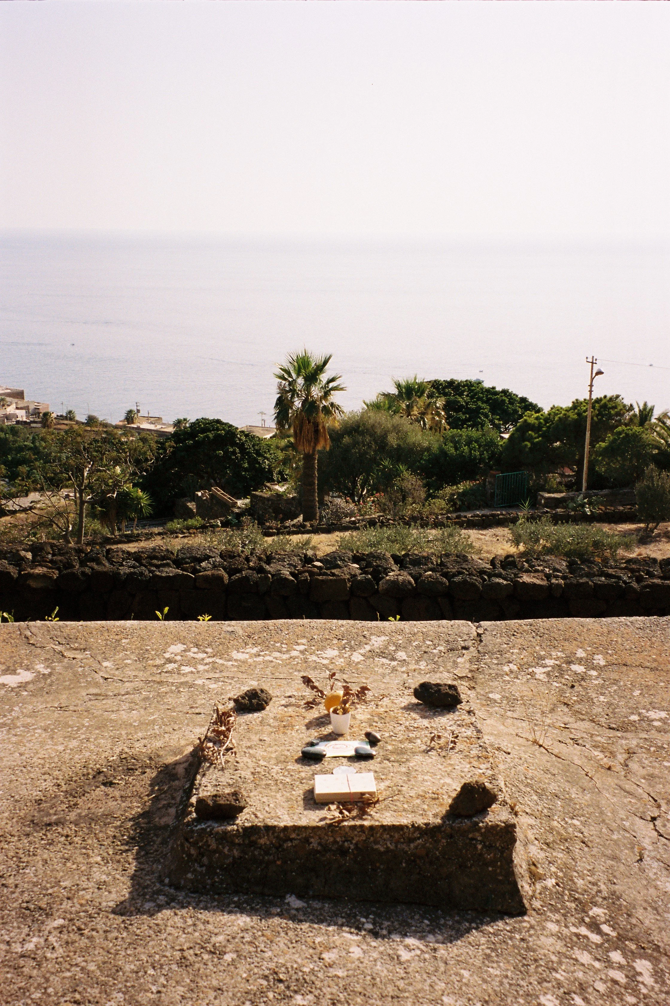 A small memorial with a cross, a candle, and flowers on a stone surface, overlooking lush green trees, palm trees, and the ocean in the distance.