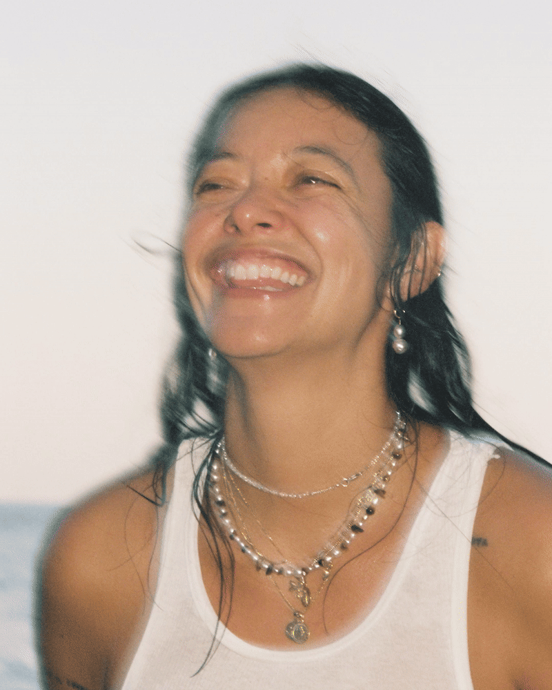 A woman with dark, wet hair wearing multiple necklaces, earrings, and a white tank top, laughing and smiling outdoors.