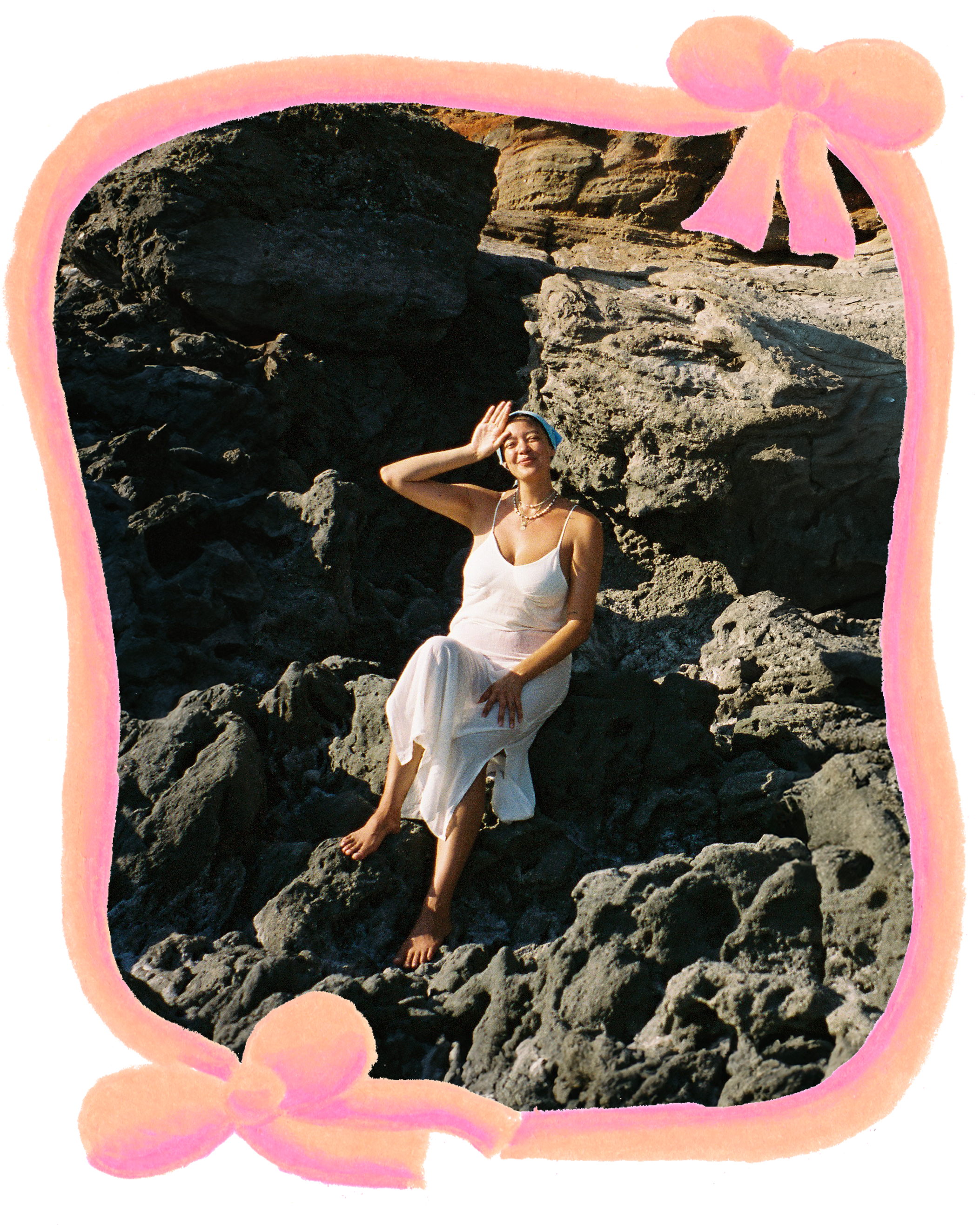 A woman in a white dress sitting barefoot on rocks at the beach, surrounded by a pink decorative frame with bows, smiling and saluting.