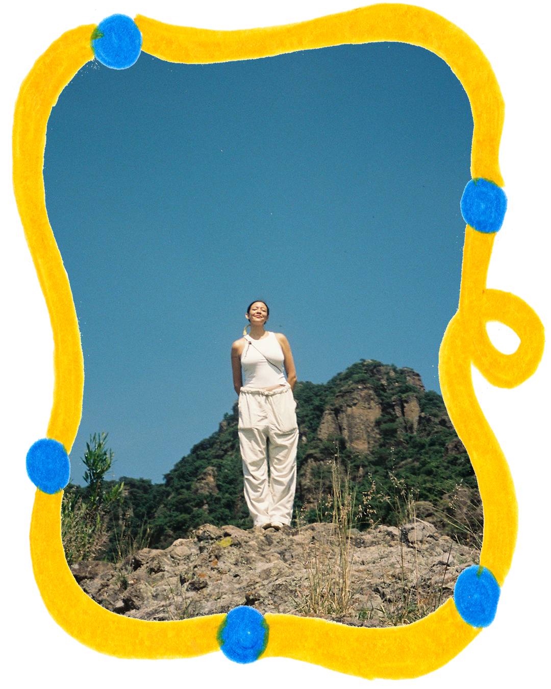 A young woman stands on rocky terrain with green hills and a clear blue sky in the background, smiling with her eyes closed.