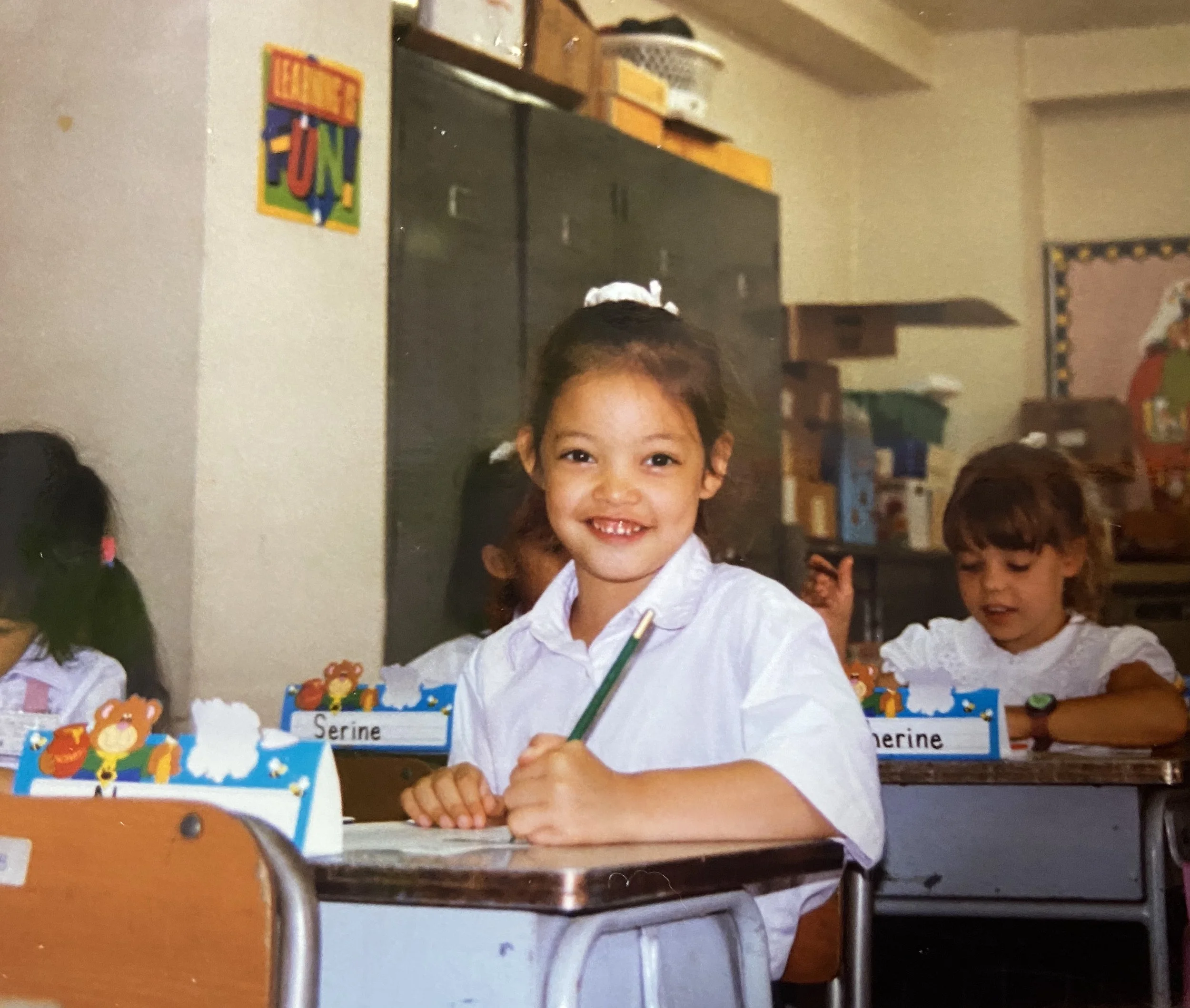 A young girl smiling brightly at her desk in a classroom, surrounded by her classmates. She has dark hair tied with white ribbons and is wearing a white shirt. The classroom has colorful decorations and a chalkboard in the background.