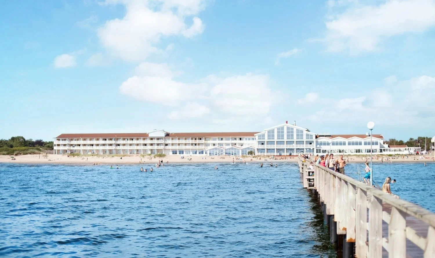 En strand med ett sockelhus i bakgrunden och en brygga som går ut i vattnet, många personer som badar och solar.