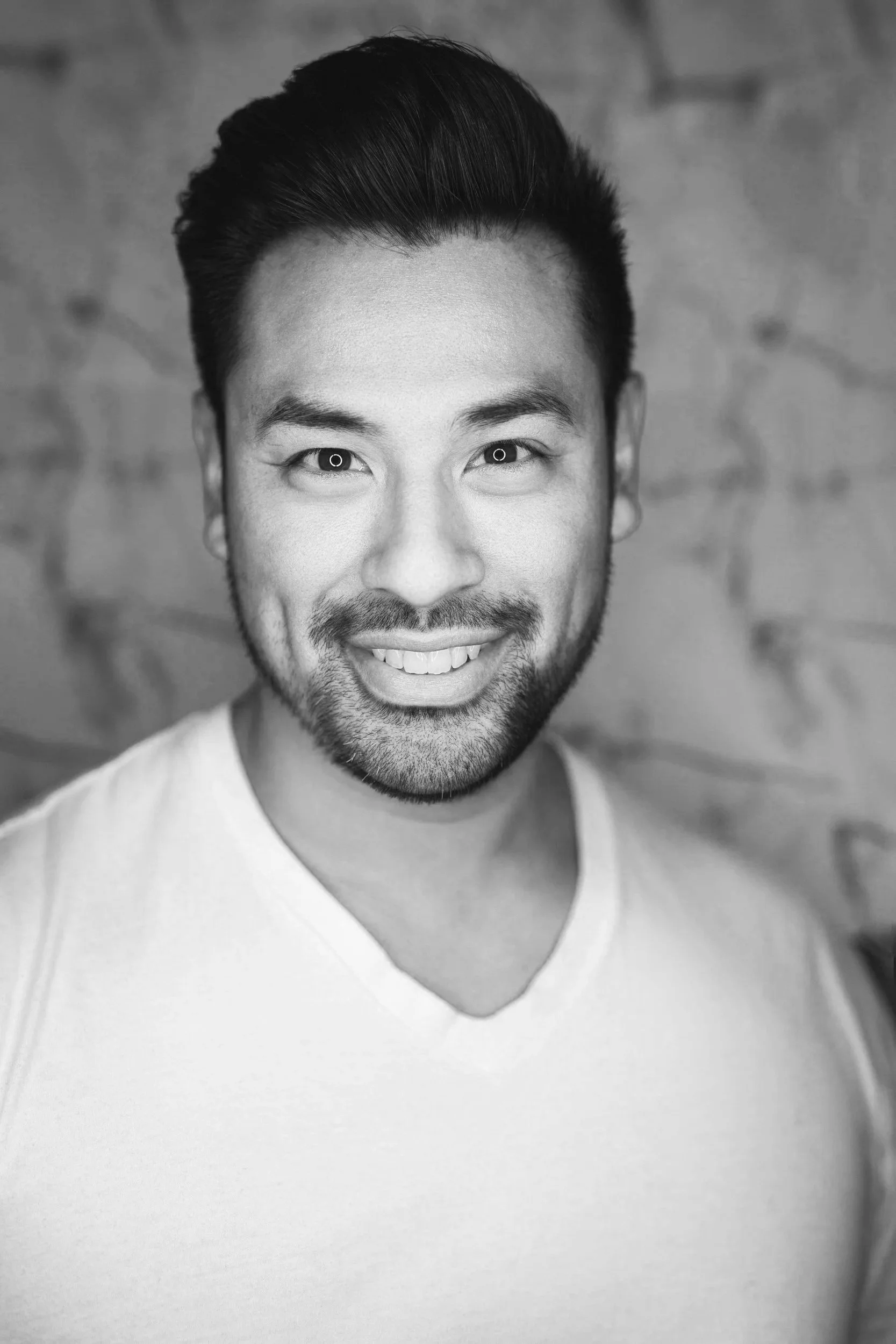 Svartvit porträttbild av en man med mörkt hår, skägg och vit T-shirt, ler mot kameran mot en tegelvägg.