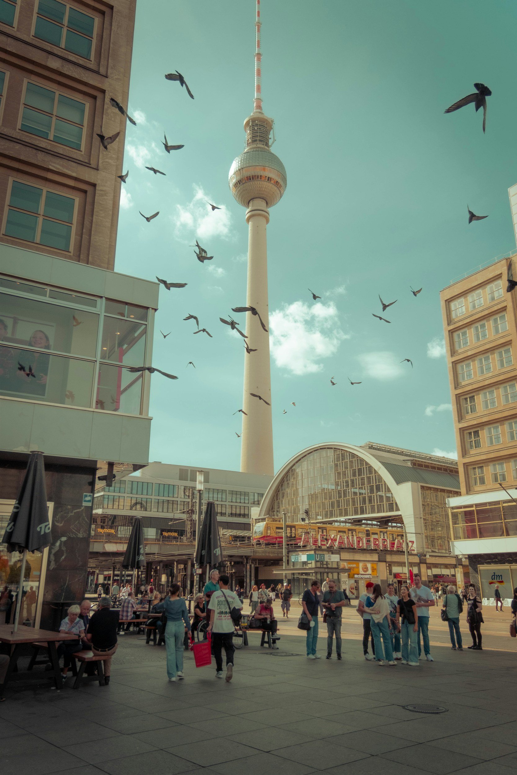Bild av Alexanderplatz i Berlin, med folk som går och sitter på uteserveringar, byggnader, och TV-tornet Alexanderplatz i bakgrunden.