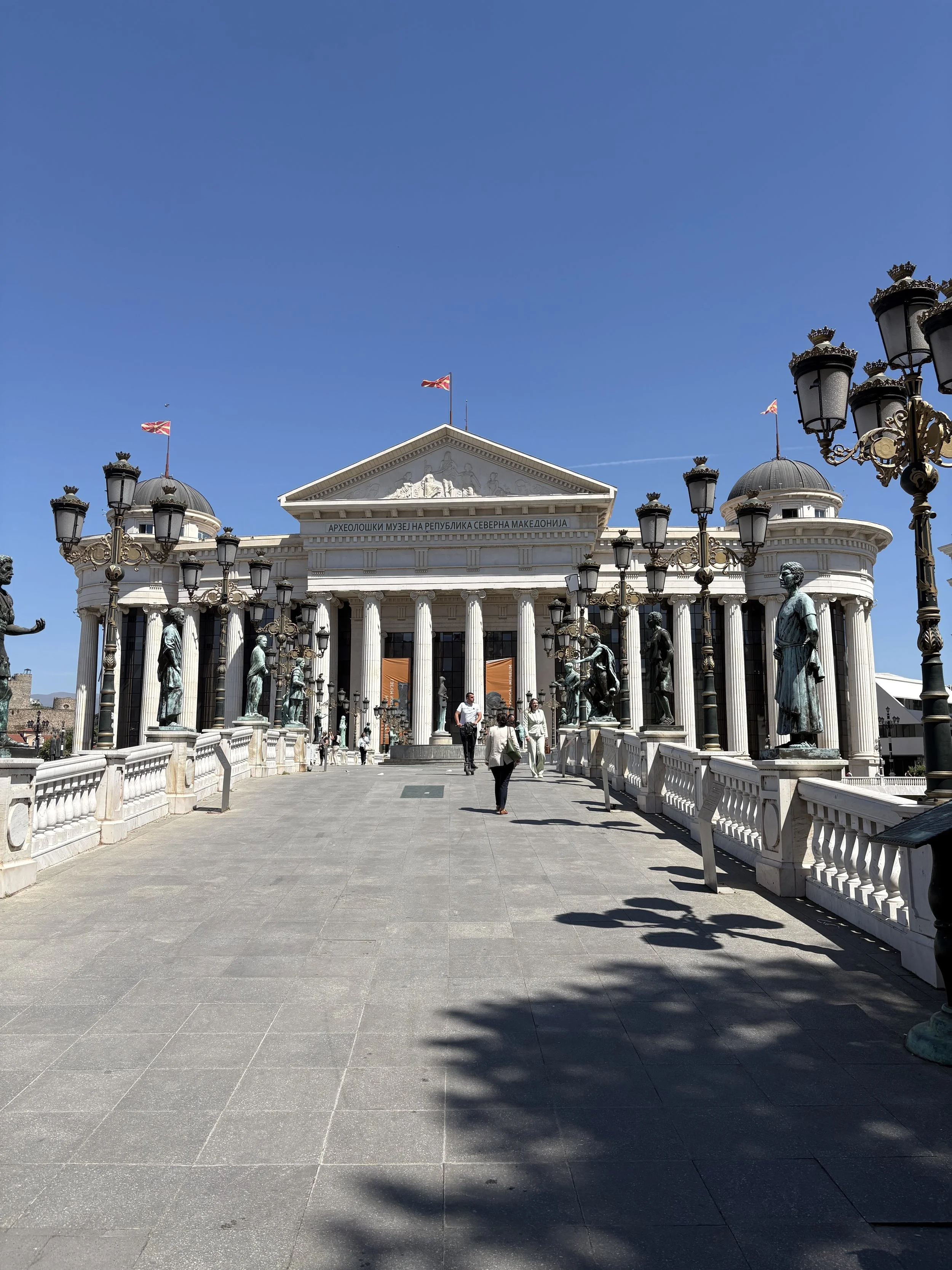 Bild av en byggnad med kolonner och statyer framför, som är arkeologimuseet i Nordmakedoniens huvudstad Skopje, med en klar blå himmel och flaggor på taket.