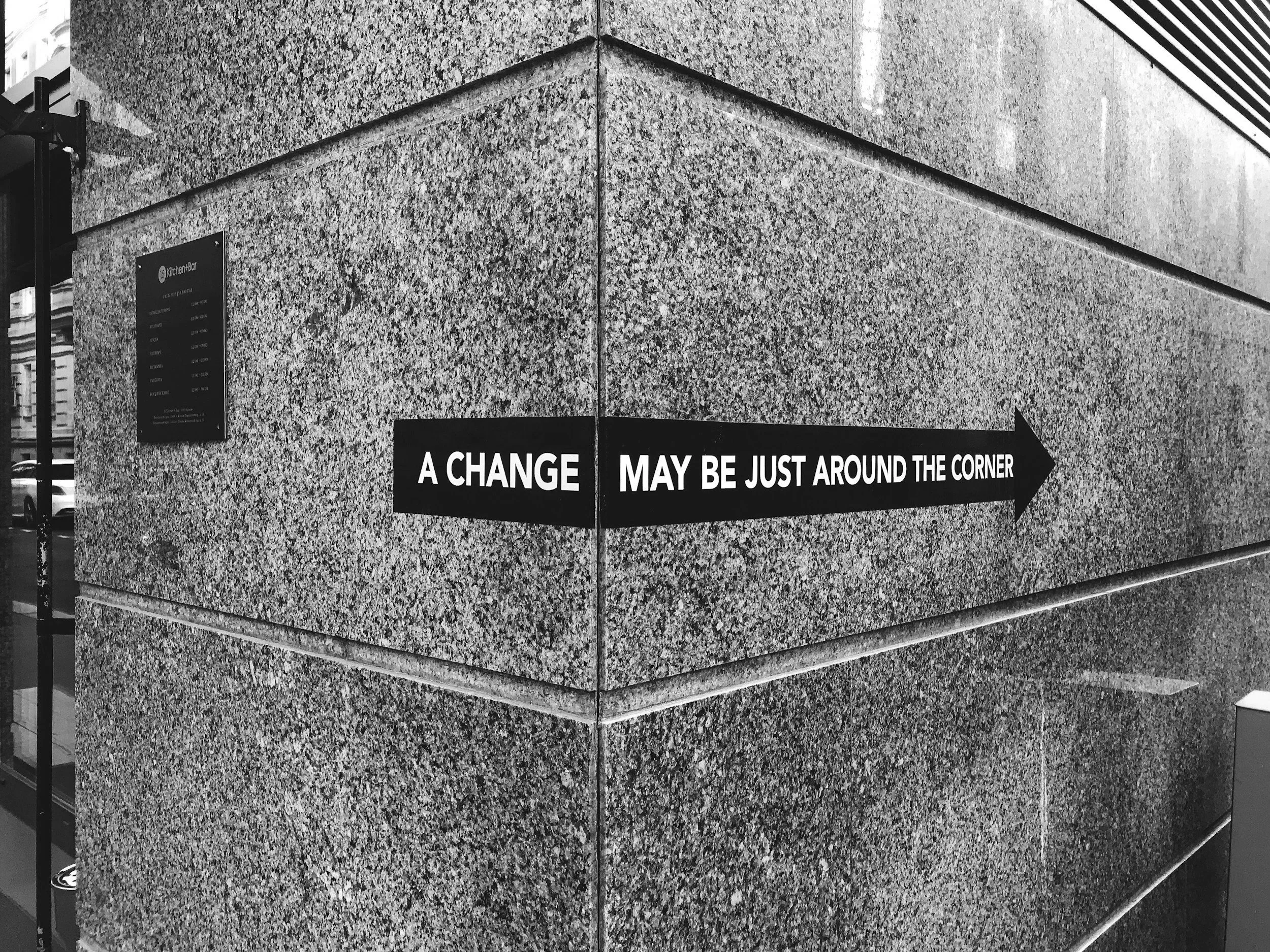 A black arrow-shaped sign on a stone wall reads, 'A CHANGE MAY BE JUST AROUND THE CORNER'.