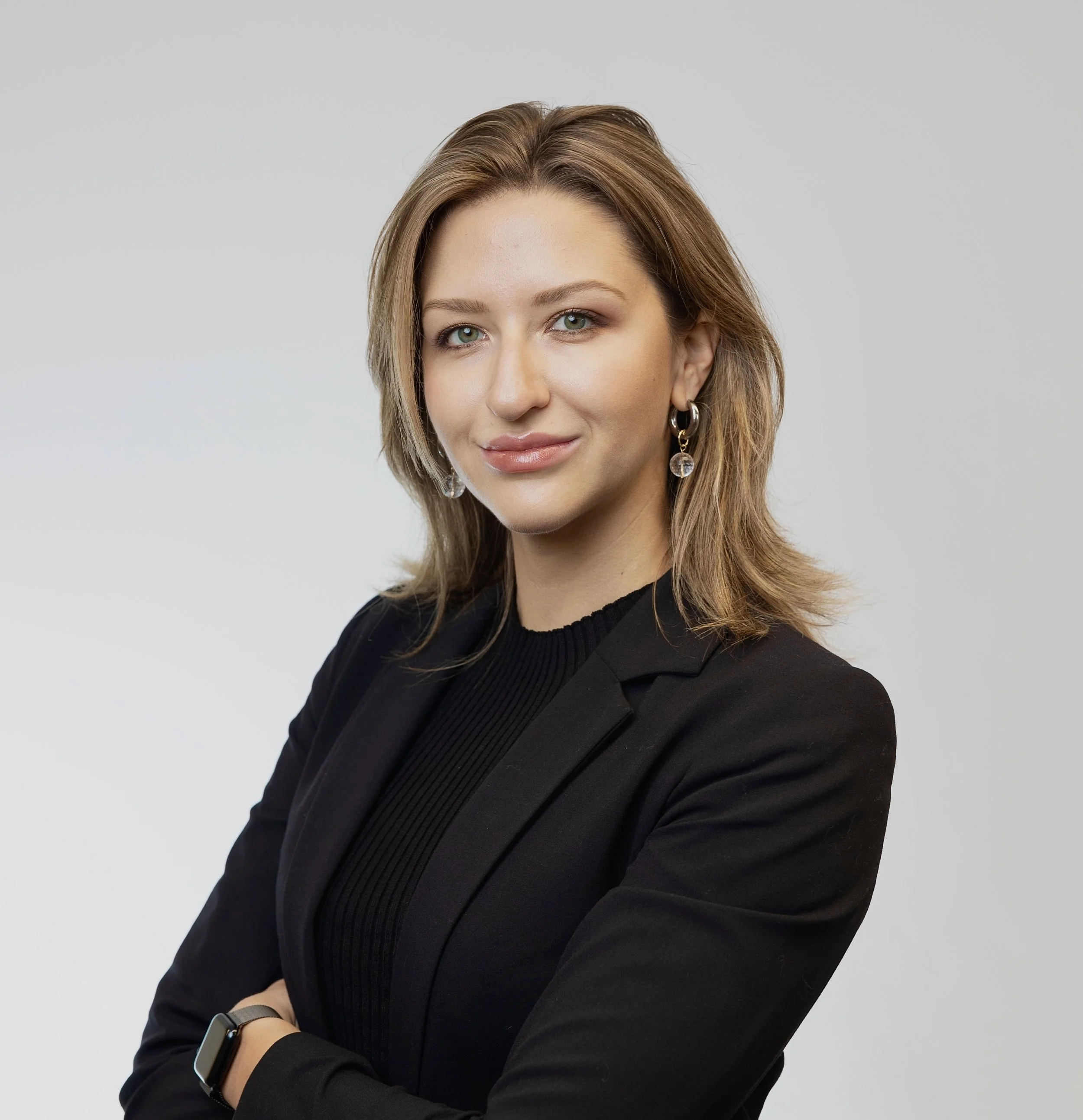 A woman with shoulder-length light brown hair, wearing a black blazer, black shirt, silver earrings, and a smartwatch, standing with arms crossed against a light grey background.