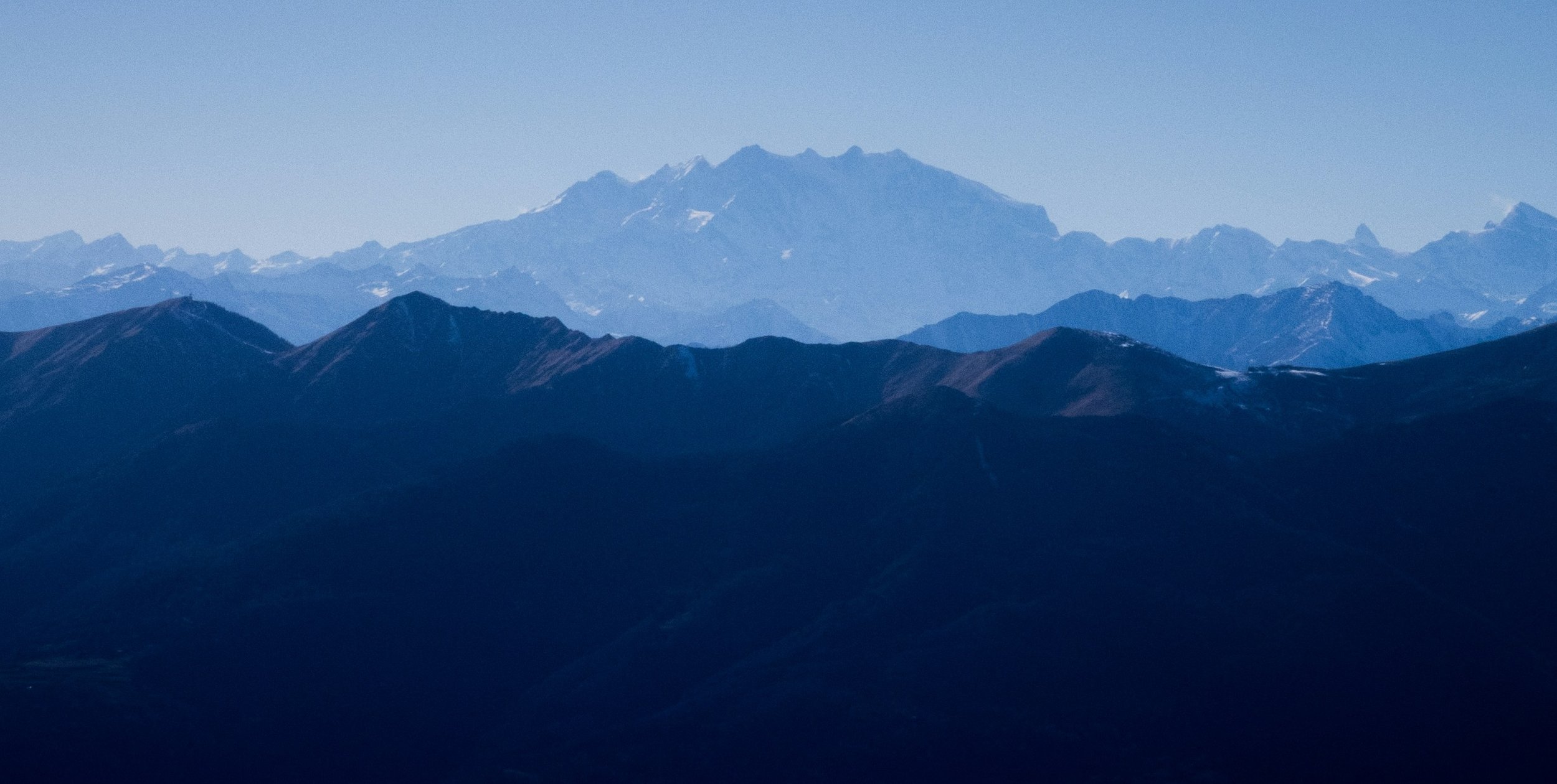 A view of layered mountain ranges with a tall, snow-capped mountain in the distance under a clear blue sky.