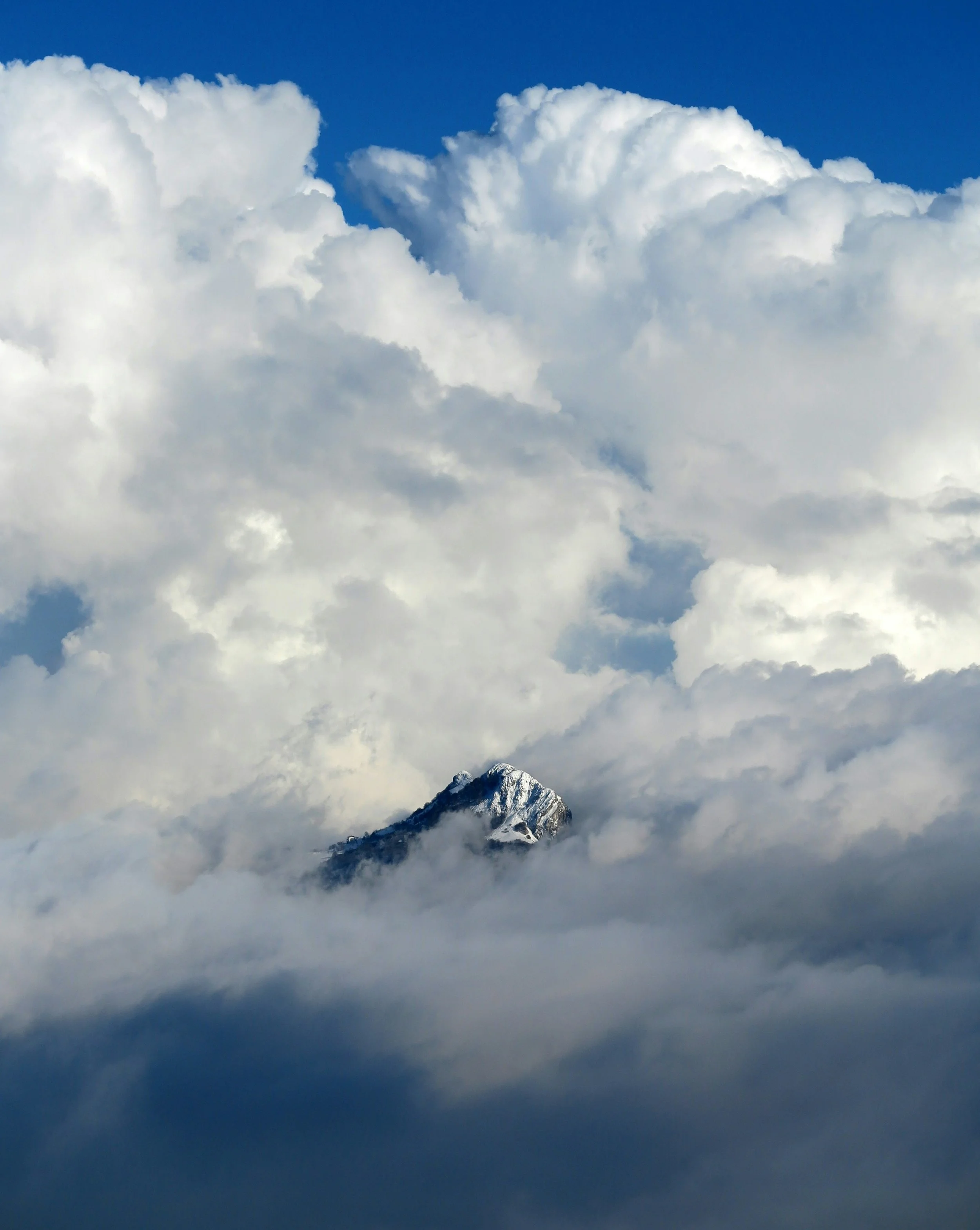 Snow-capped mountain peak emerging through clouds under a blue sky with large white clouds.