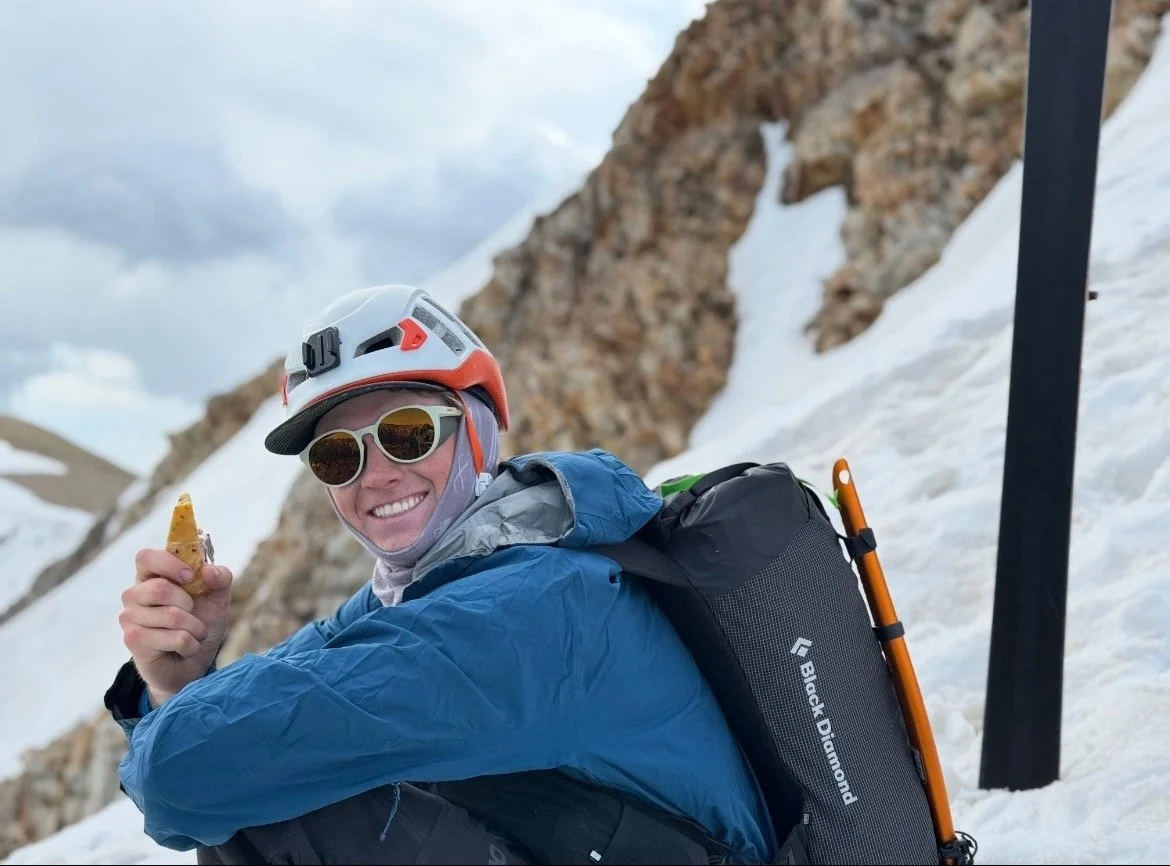 Young woman with curly hair, glasses, and a bright smile in an indoor setting.