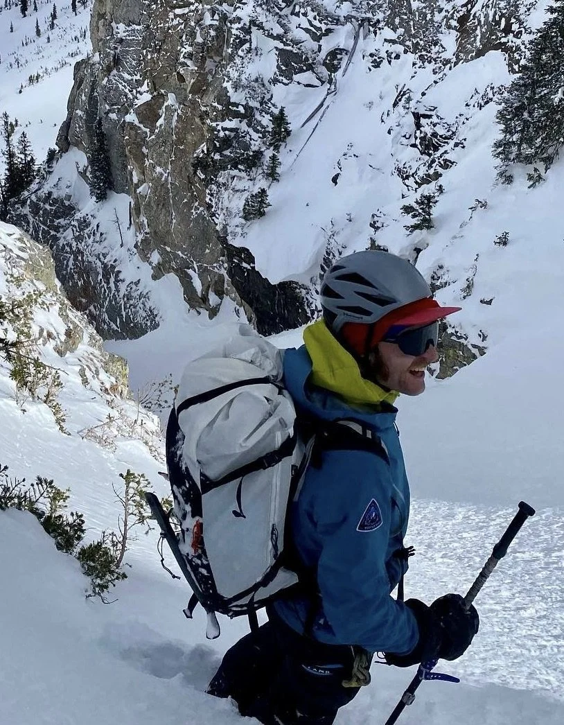 A man with a helmet, sunglasses, and a mustache smiling in a snowy mountain landscape.