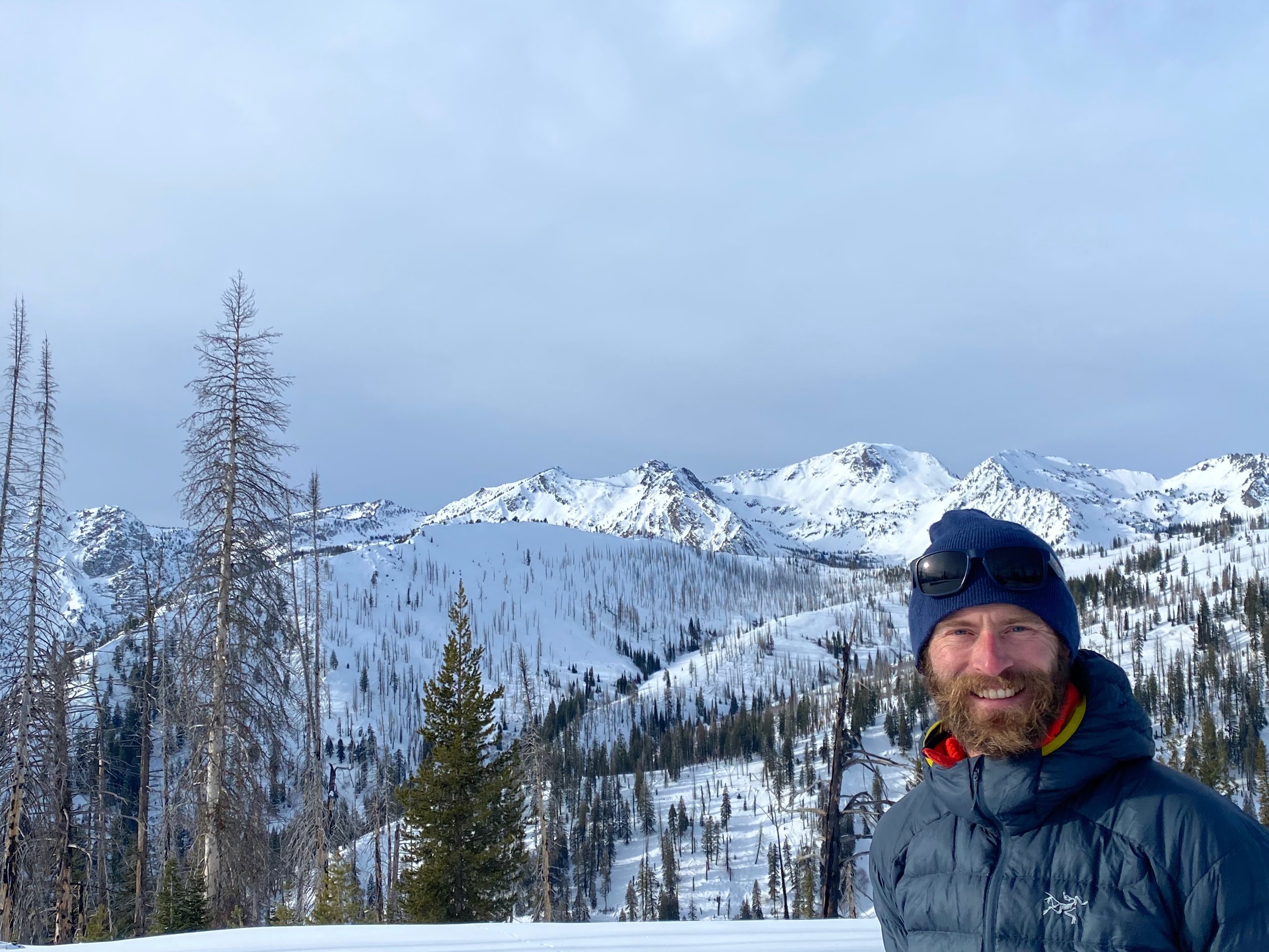 Man with a beard smiling outdoors near a lake, wearing a blue beanie and a brown jacket with yellow zipper, with a forest and blue sky with clouds in the background.