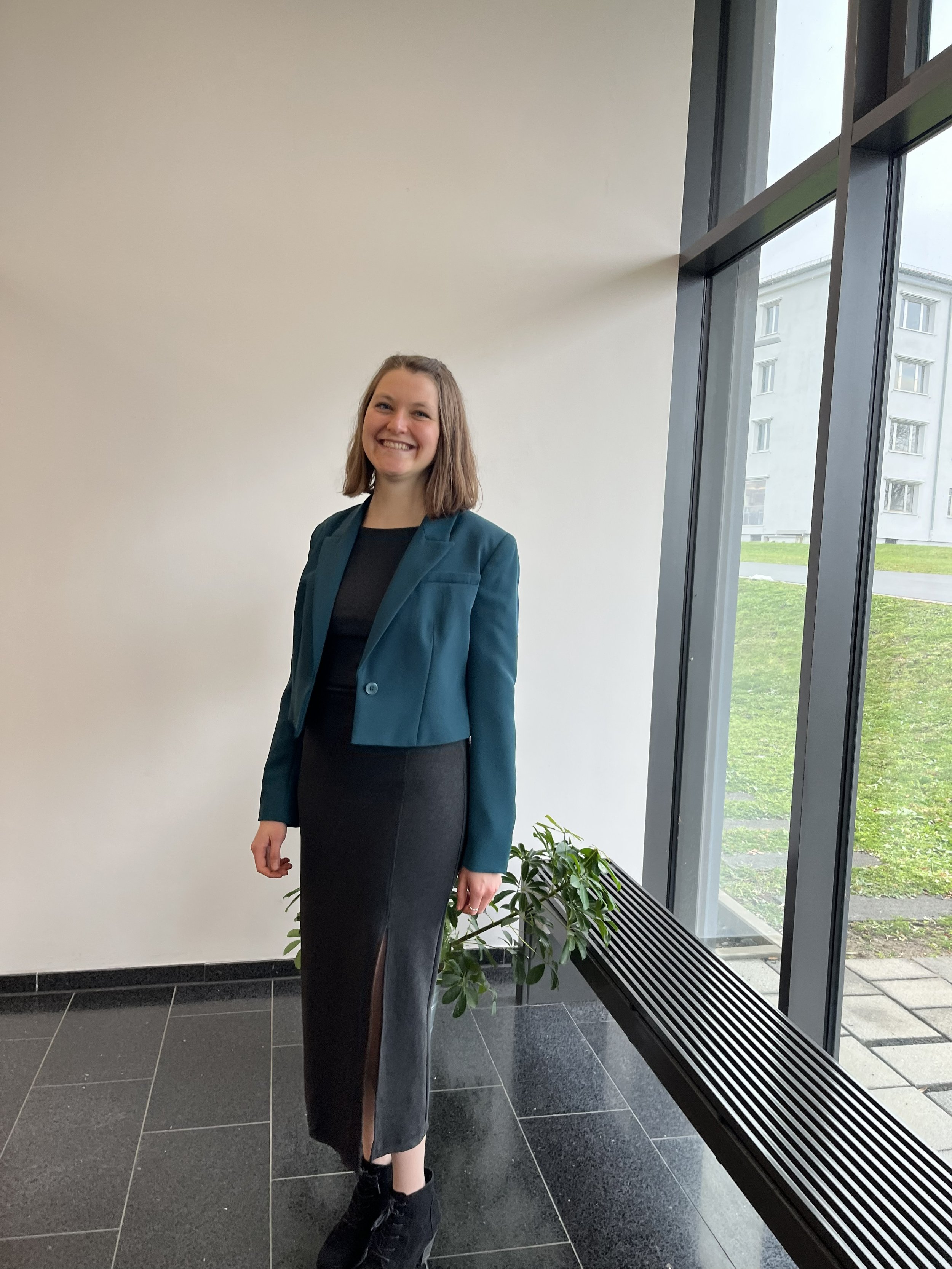 Junge Frau in Anzug steht neben Fenster mit Blick nach draußen, im Hintergrund grüne Wiese und Gebäude.