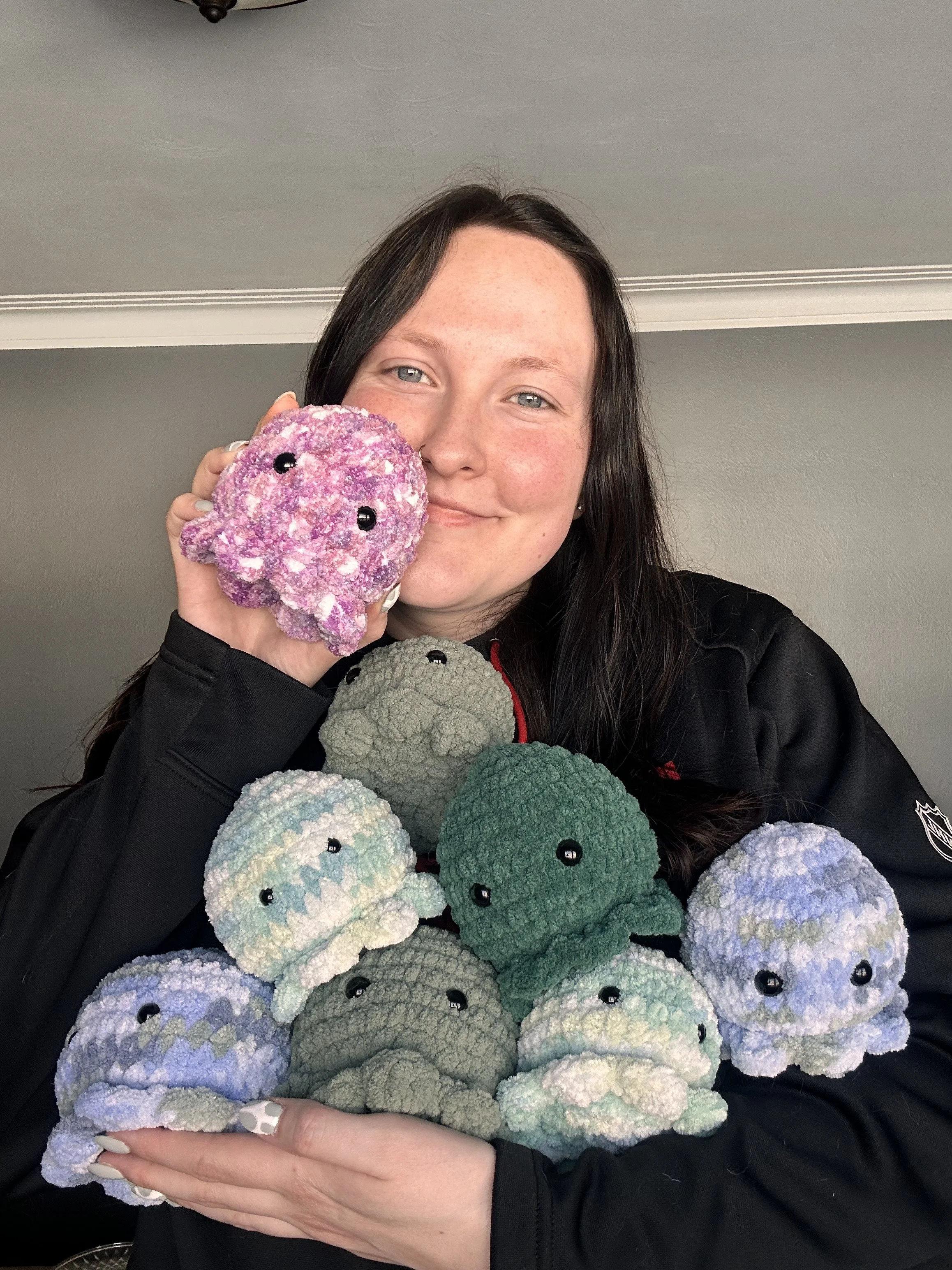 A woman with long dark hair and light skin holding a plush octopus toy with colorful, textured fabric and black eyes. Multiple stuffed octopus toys in various colors are visible in front of her.