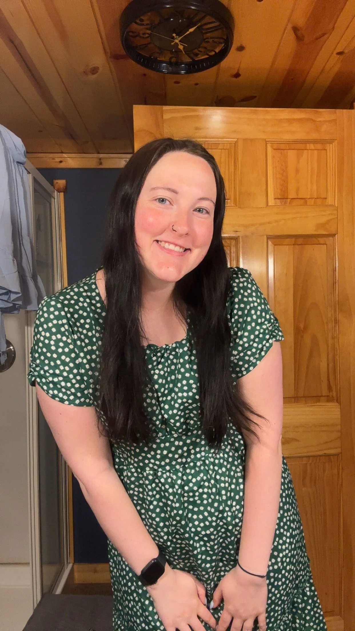 Smiling woman with long dark hair, wearing a green dress with white polka dots, standing indoors in front of a wooden door and wall, with a black wall clock on the ceiling above her.