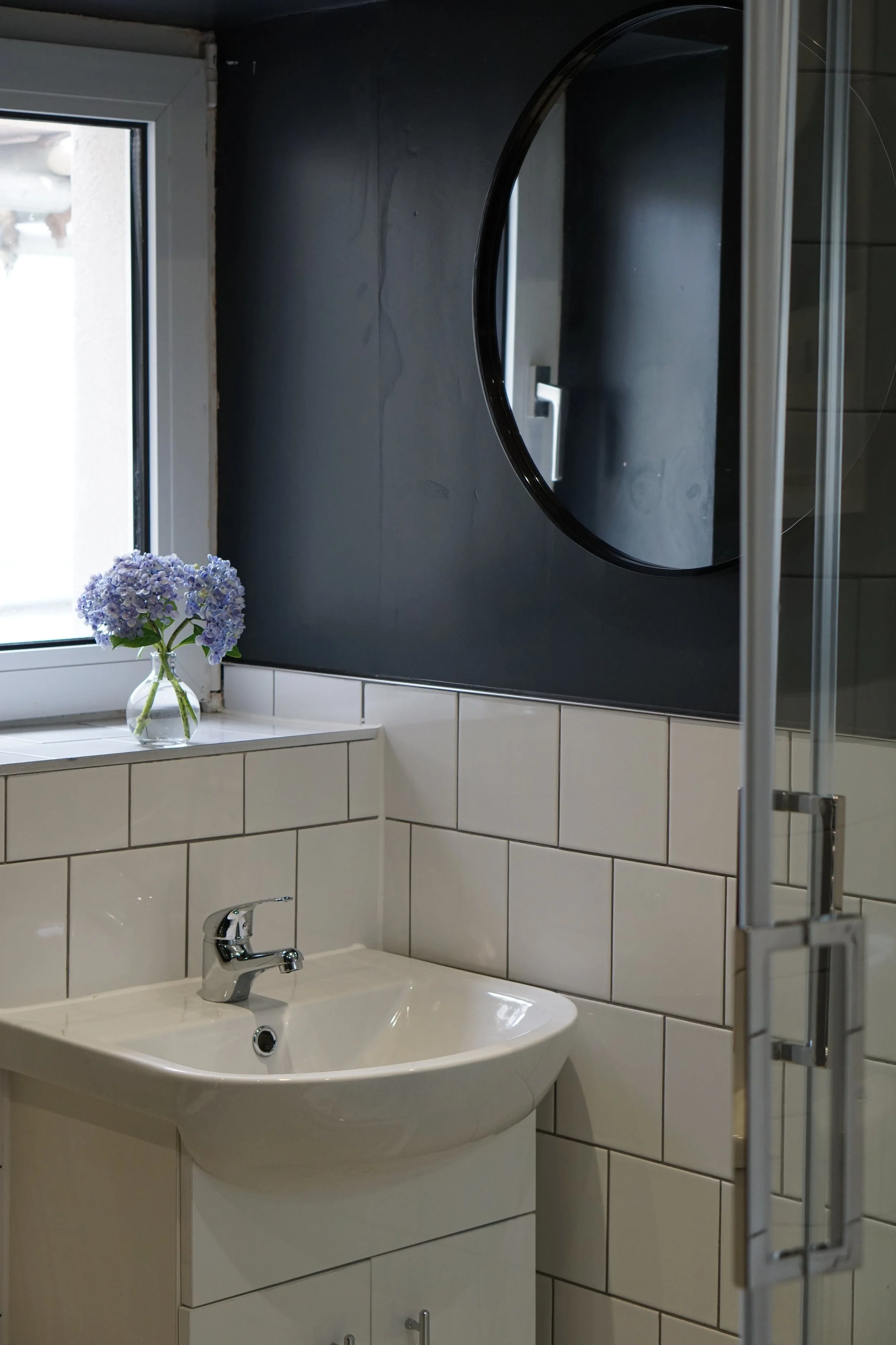 Bathroom with a white sink, a round mirror, and a vase of purple hydrangeas on the windowsill.