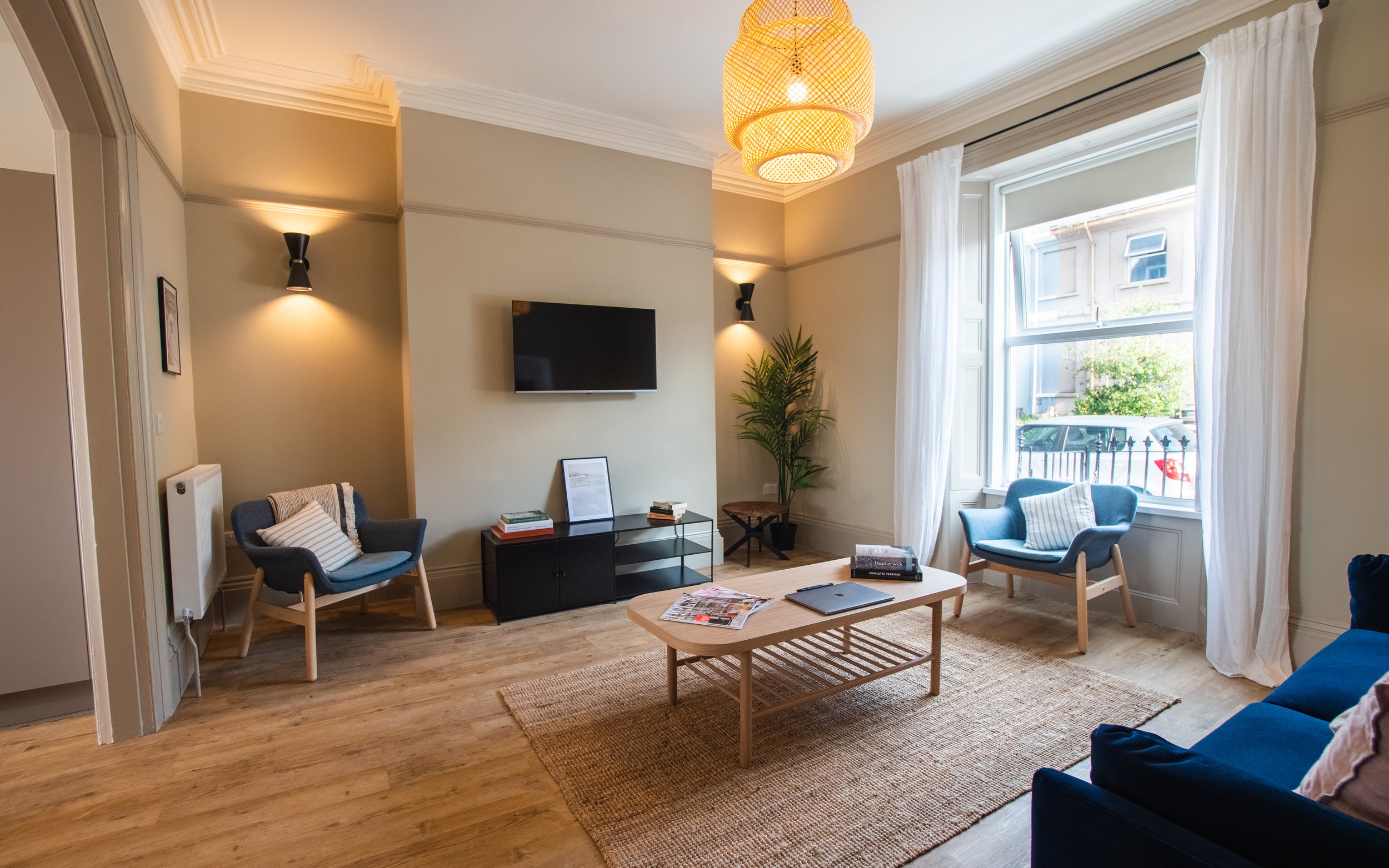 Bright living room with a large window, blue armchairs, a coffee table with magazines and a laptop, a television on the wall, and a plant near the window.