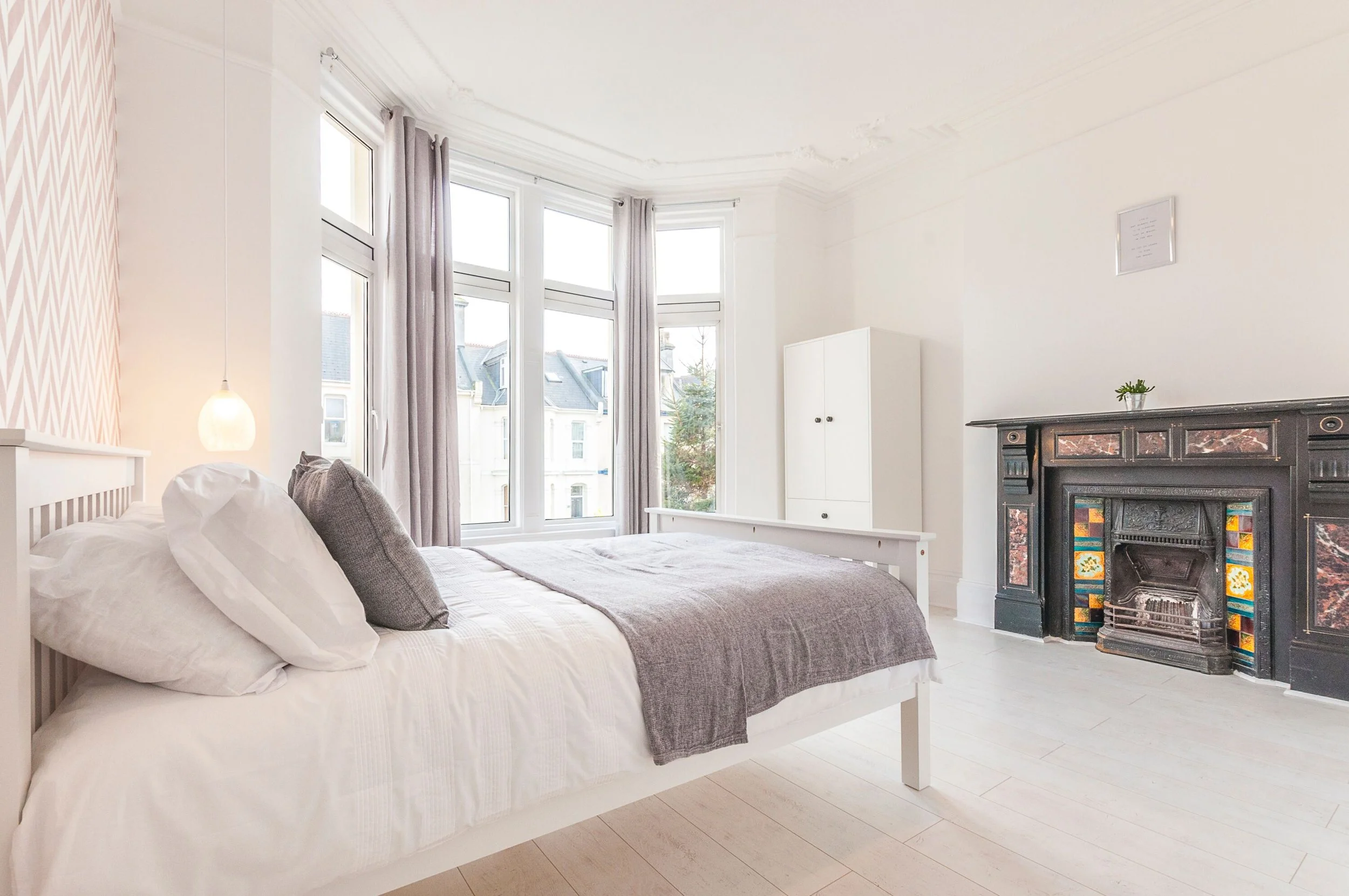 Bright bedroom with large bay window, white bed with gray blanket and pillows, white wardrobe, decorative black fireplace with colorful tiles, small potted plant on fireplace mantel, framed art on white wall, light wood flooring, and gray curtains.