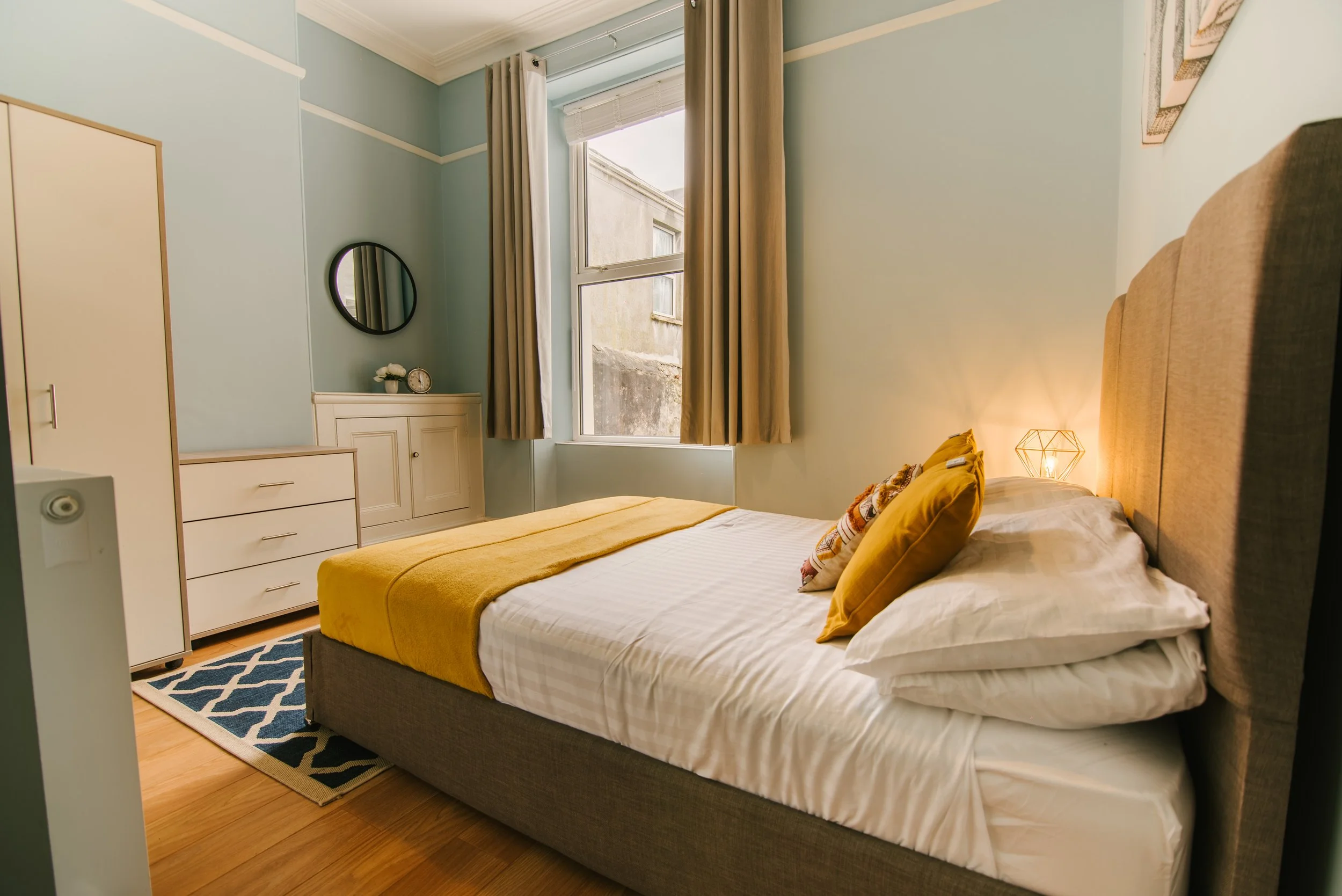 A cozy bedroom with a large bed featuring a beige upholstered headboard, yellow pillows, and a yellow blanket. The room has light blue walls, a window with beige curtains, and a small blue and white geometric rug. There is a white dresser, a cabinet 