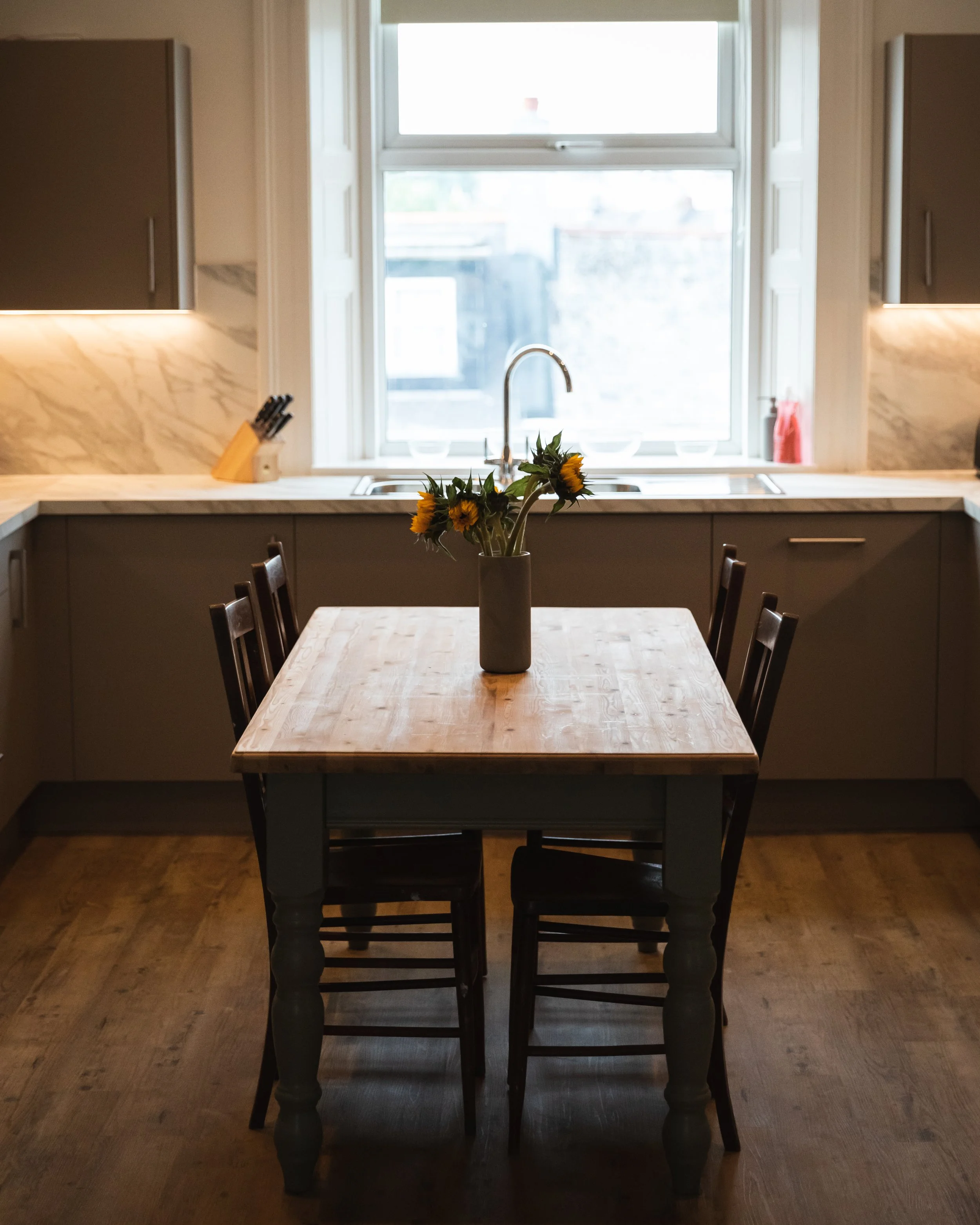 A wooden dining table with a vase of sunflowers in the center, surrounded by six dark wooden chairs, in a kitchen with a window and beige cabinets.