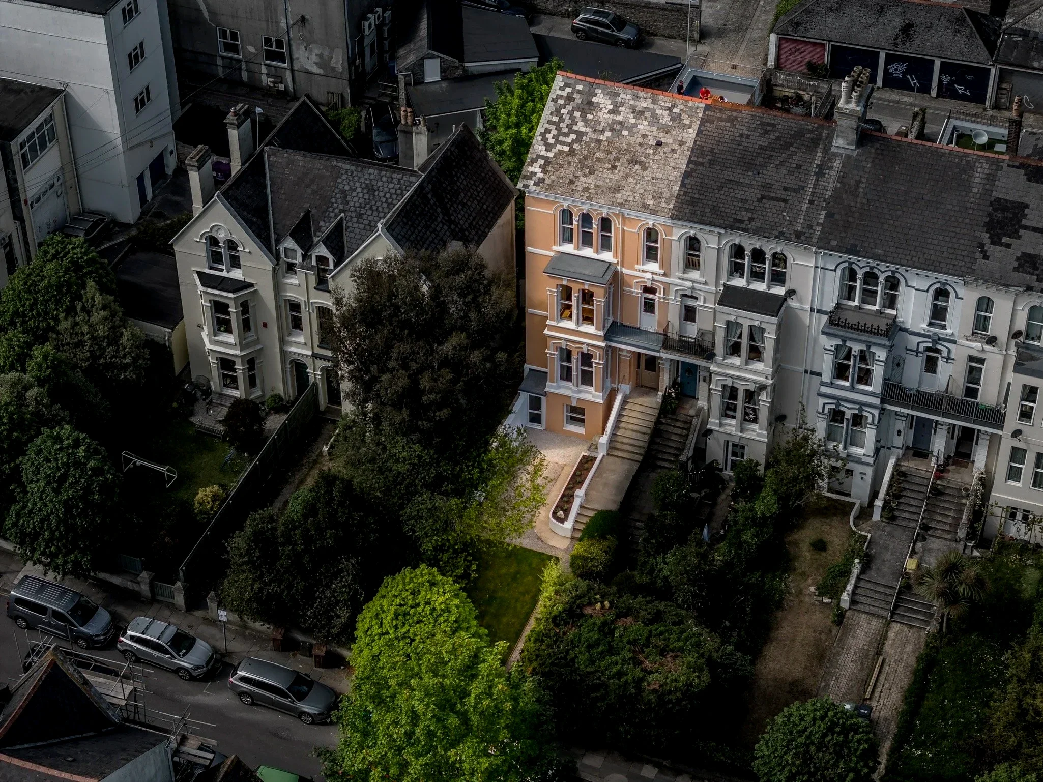 Aerial view of a city neighborhood with three multi-story residential buildings, trees, parked cars, and narrow streets.