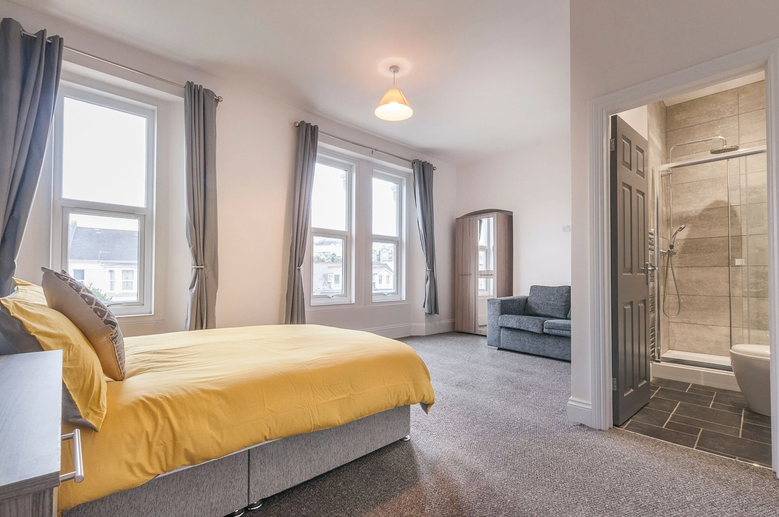 A bedroom with a yellow bedspread, gray pillows, and large windows with gray curtains. There is a gray sofa, a wooden wardrobe, and a small doorway leading to a bathroom with a shower and toilet.