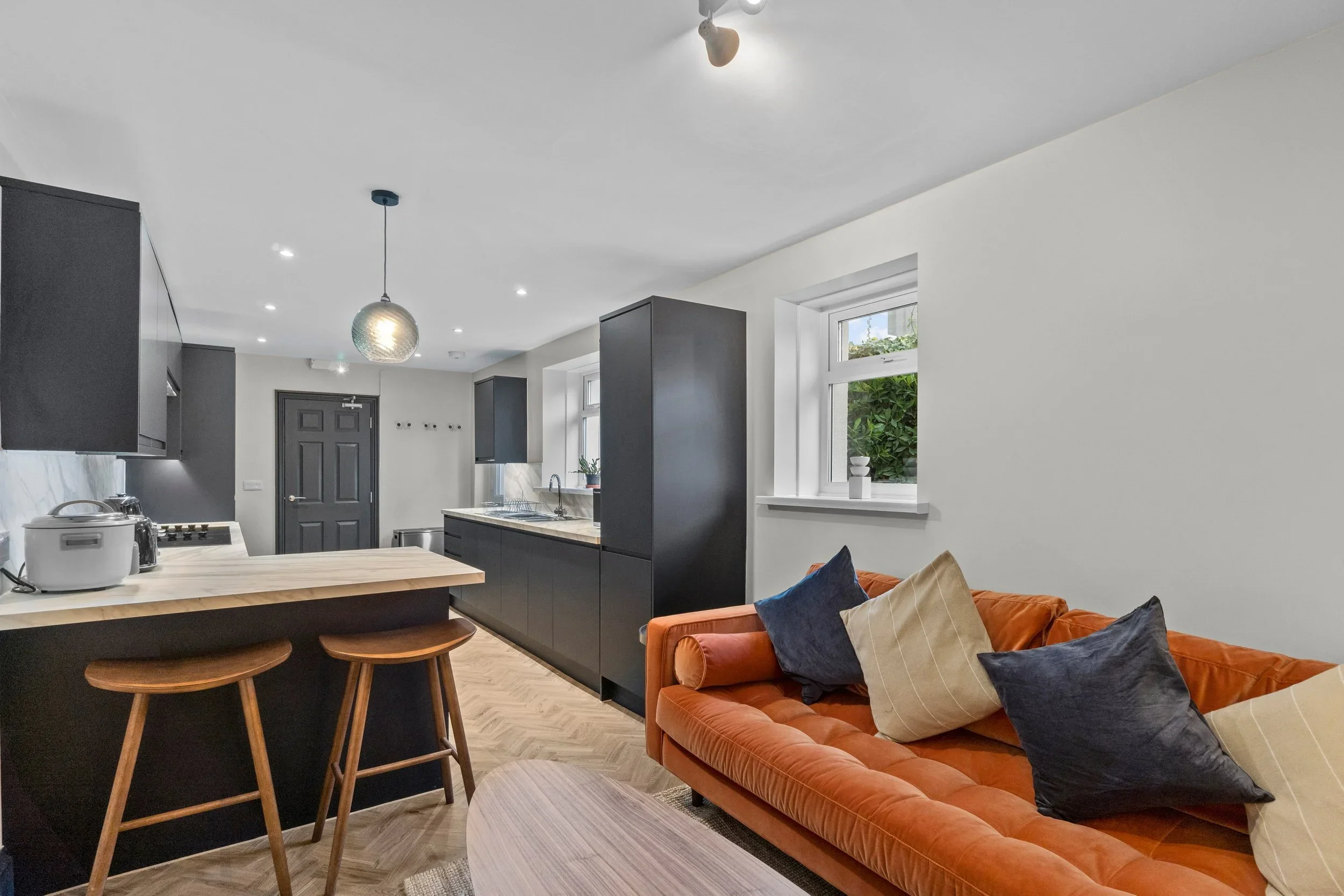 Open-concept living room and kitchen with black cabinetry, an orange velvet sofa with throw pillows, and a window with greenery outside.