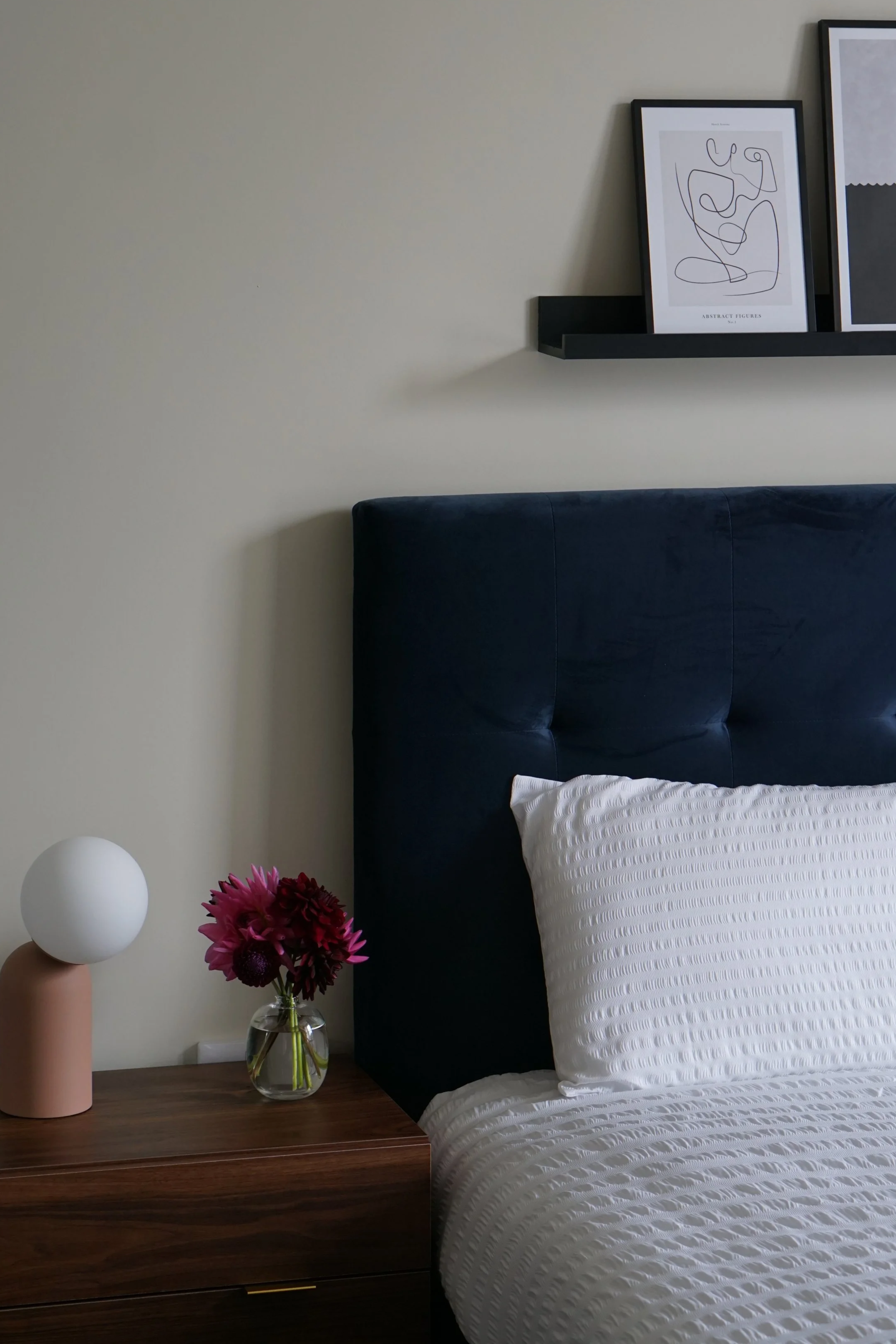 A bedroom with a dark navy upholstered headboard, a white textured pillow, a wooden nightstand with a small vase of pink and purple flowers, a modern lamp with a beige base and white spherical shade, a black floating shelf with framed abstract line a