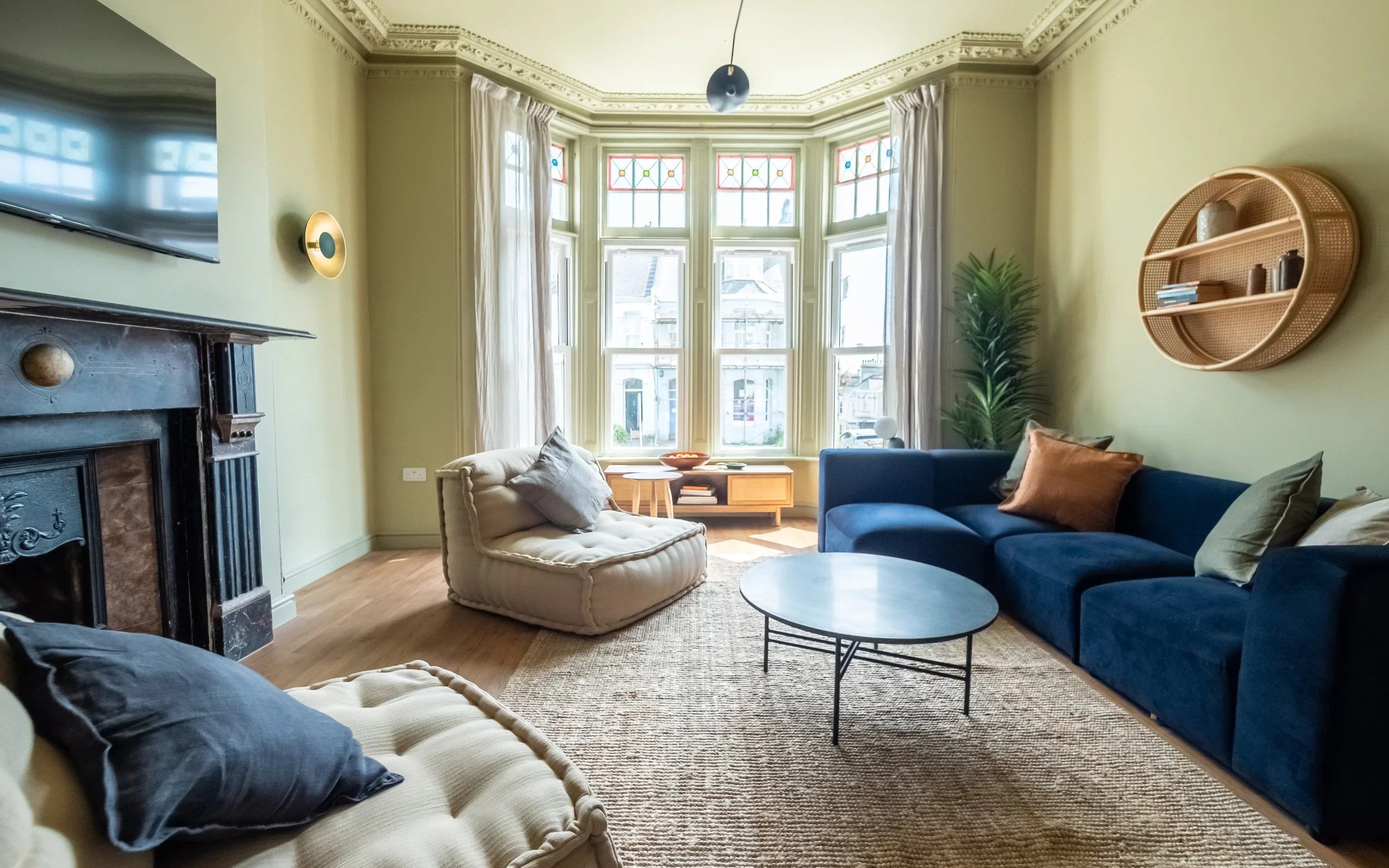 Living room with large bay window, navy blue sofa, beige armchair, marble coffee table, black fireplace, wall-mounted TV, woven wall shelf, and green plants.