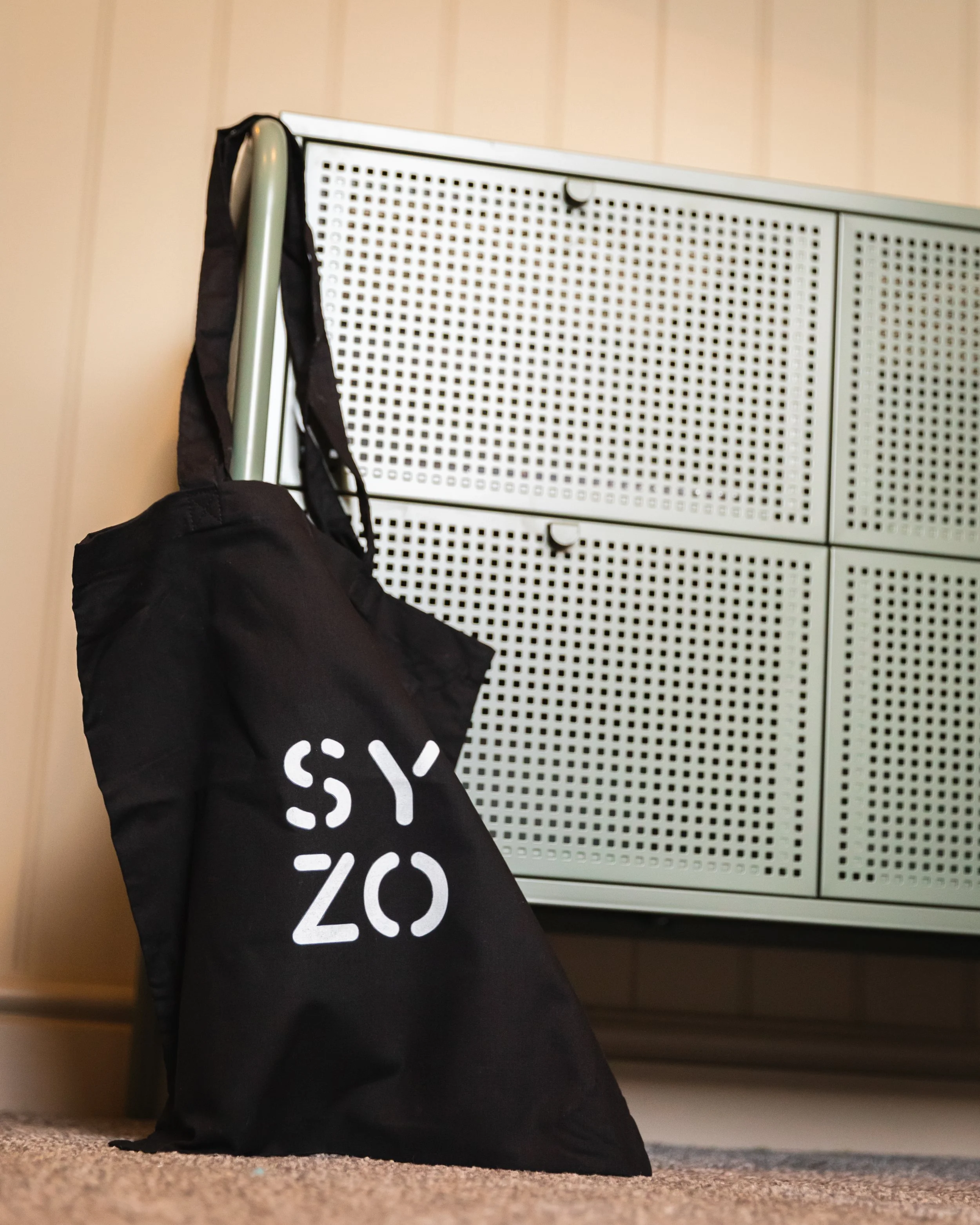 A black tote bag with white text leaning against a metal shelf with perforated doors, set against a wood-paneled wall.