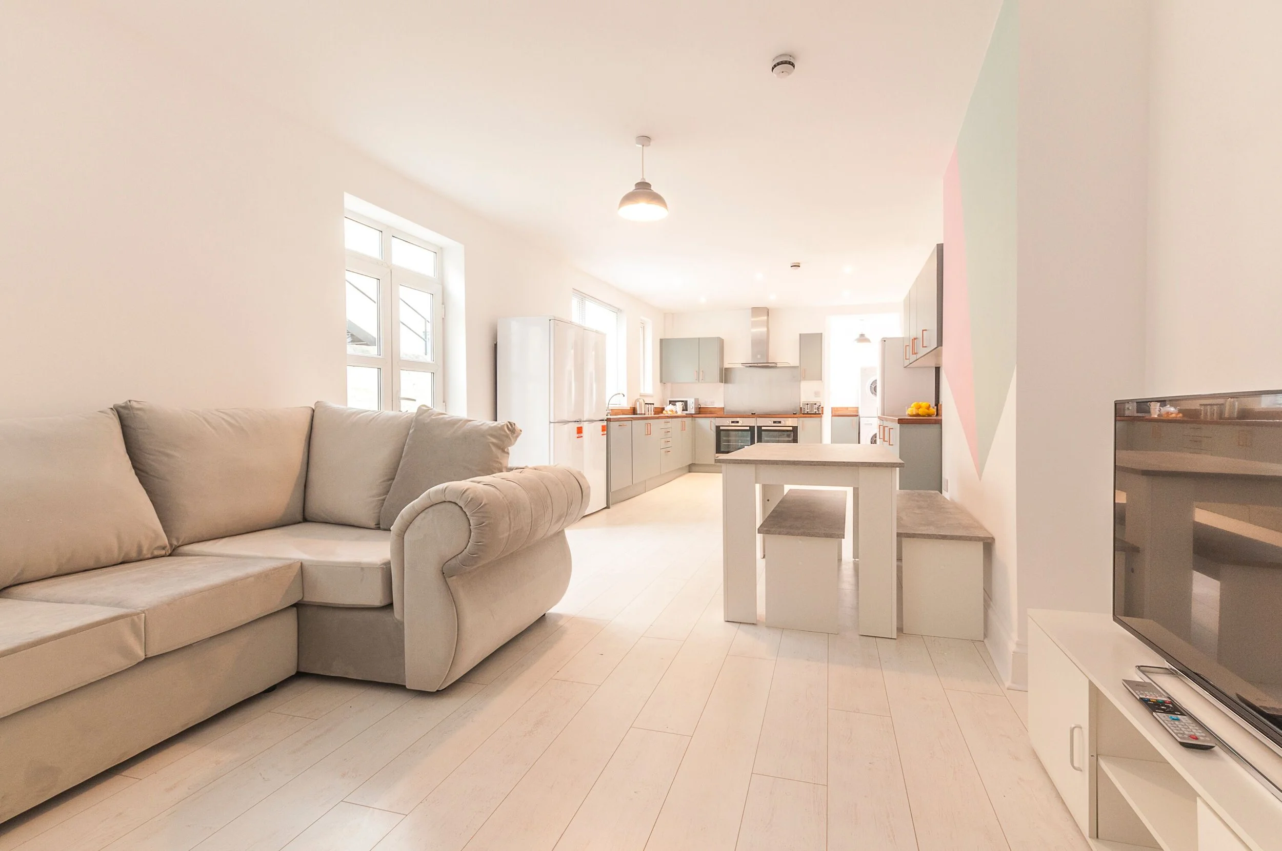Bright, open-concept living room and kitchen with white walls, light wood flooring, beige couch, white dining table, gray cabinets, and stainless steel appliances.