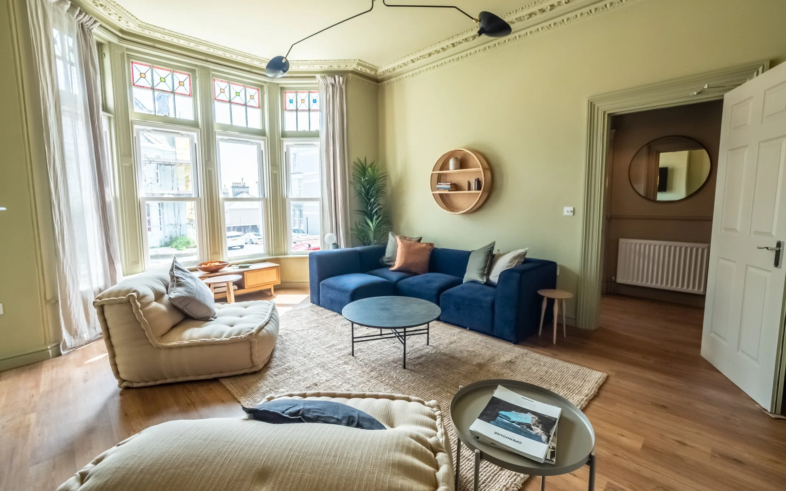 Living room with large bay window, blue sofa, beige armchairs, round coffee table, small side table, wall shelf, potted plant, and decorative trim on the ceiling.