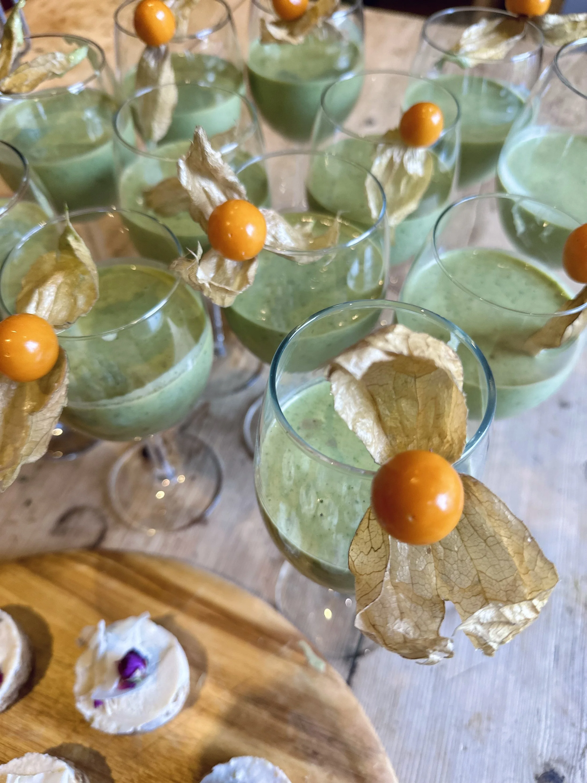 Several glasses of green mousse topped with a physalis fruit and a leaf, set on a wooden table.