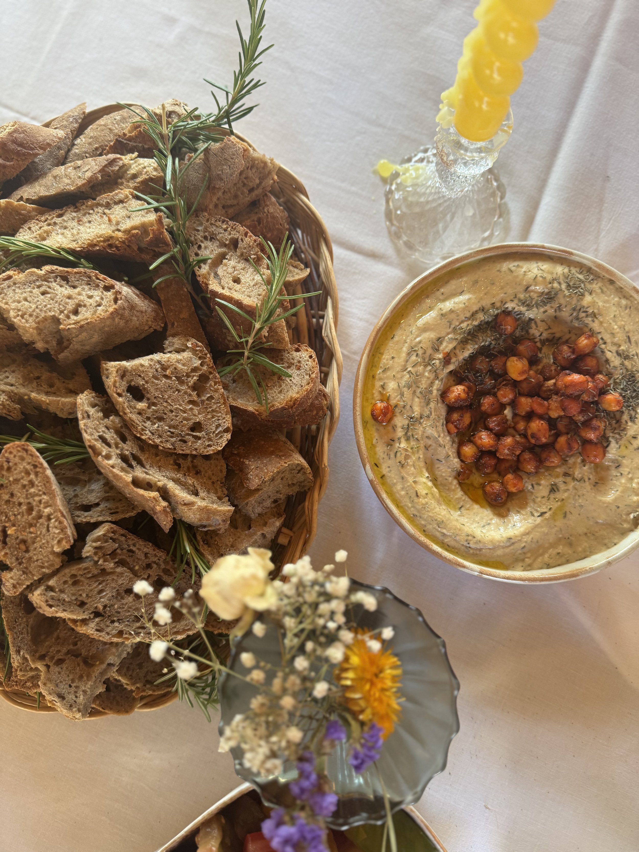 Basket of sliced bread with sprigs of fresh rosemary, bowl of hummus topped with roasted chickpeas and herbs, small vase of colorful flowers, and a lit yellow beeswax candle on a white tablecloth.