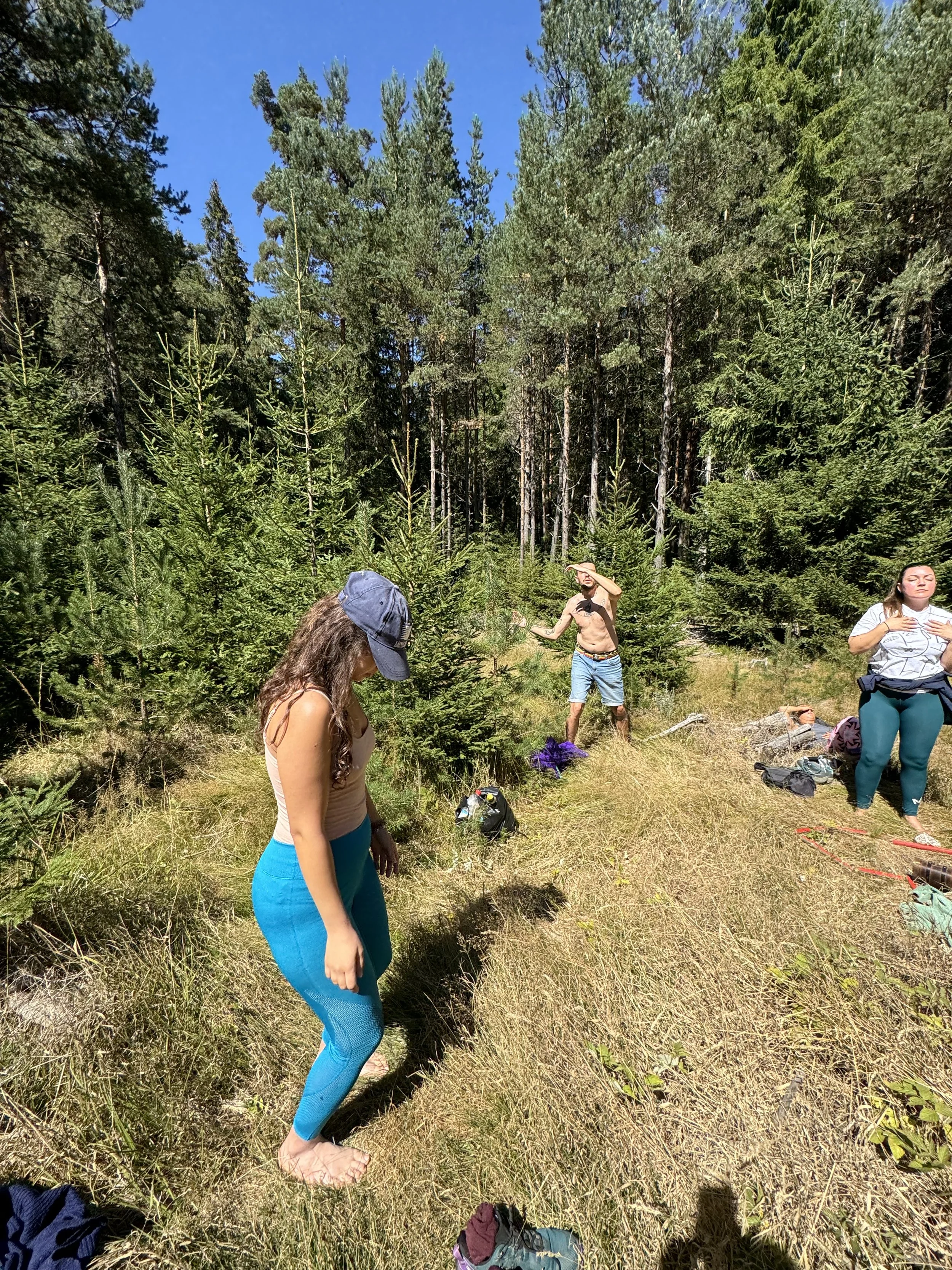 Group of people outdoors in a forest, some standing and some sitting, with backpacks and camping gear on the ground, surrounded by tall green trees under a clear blue sky.