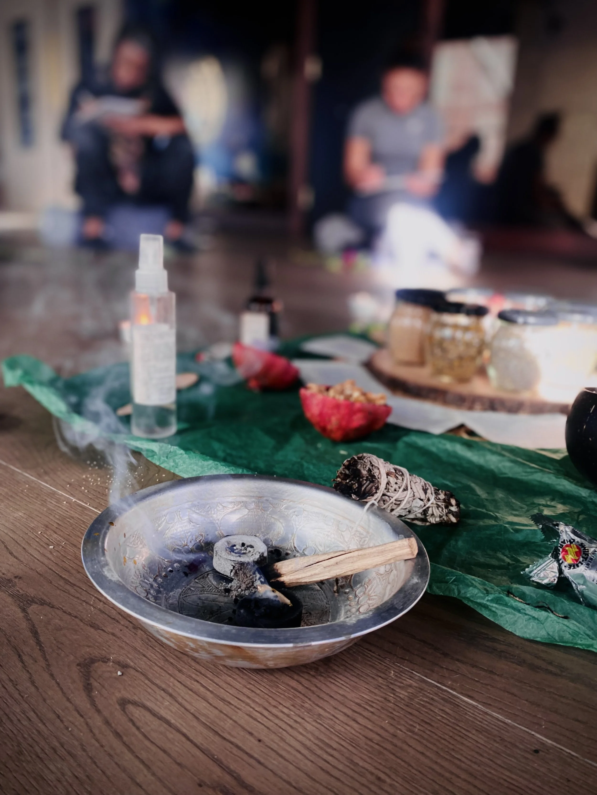A burning incense cone in a decorative metal bowl on a wooden table, with various small objects and food items in the background, and two blurred people sitting on the floor in an indoor setting.